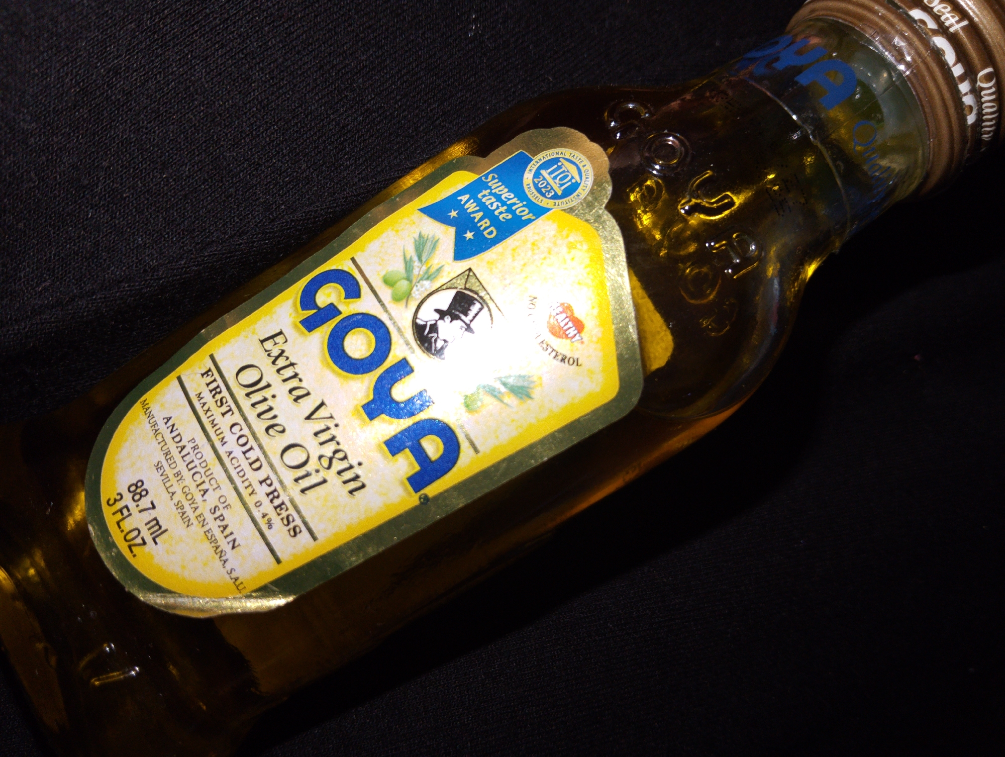 A clear glass bottle of Goya Extra Virgin Olive Oil rests on a dark fabric surface. The bottle is angled, with the label facing upwards. The label is predominantly yellow with blue and white text and graphics. It features the brand name "GOYA" in large blue letters, along with descriptions such as "Extra Virgin Olive Oil," "FIRST COLD PRESS," and "MAXIMUM ACIDITY 0.4%." Below this, it states "PRODUCT OF ANDALUCIA, SPAIN," and "MANUFACTURED BY: GOYA EN ESPAÑA, S.A.U. SEVILLA, SPAIN." There is also a blue ribbon with "Superior Taste AWARD" written on it, and a circular seal with "INTERNATIONAL TASTE & QUALITY INSTITUTE BRUSSELS 2023." The bottle contains a golden-yellow liquid, which is olive oil. Reflections are visible on the surface of the bottle. The background is a dark, textured fabric. There are no people or animals present in the image, nor any indication of the city of Lagos, Nigeria.