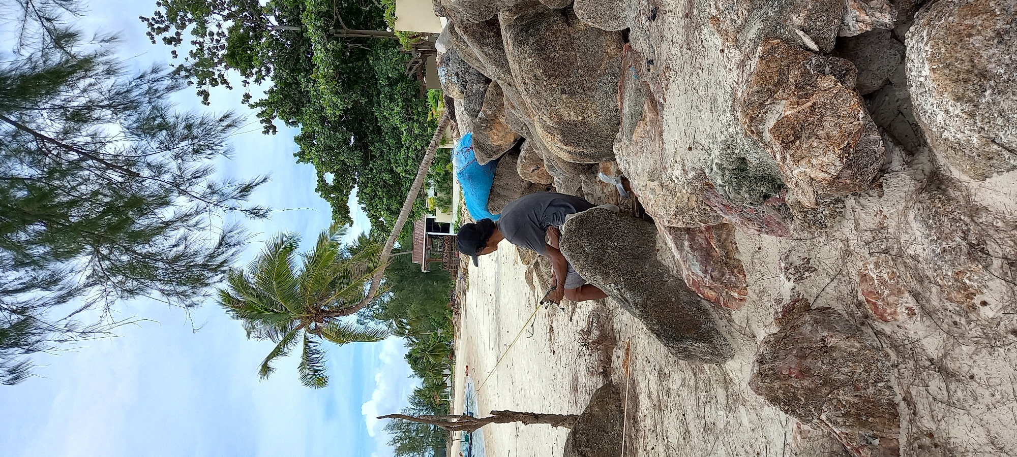 The image shows a man crouching amongst rocks at Chaloklum Beach in Baan Chaloklum, Thailand. The man wears a dark shirt, light-colored shorts, and a dark baseball cap. He is holding an object in his hands and appears to be working on something between the rocks. The beach is visible in the background, with sand and trees. A blue bag is wedged between some rocks behind the man. The sky is partly cloudy.
