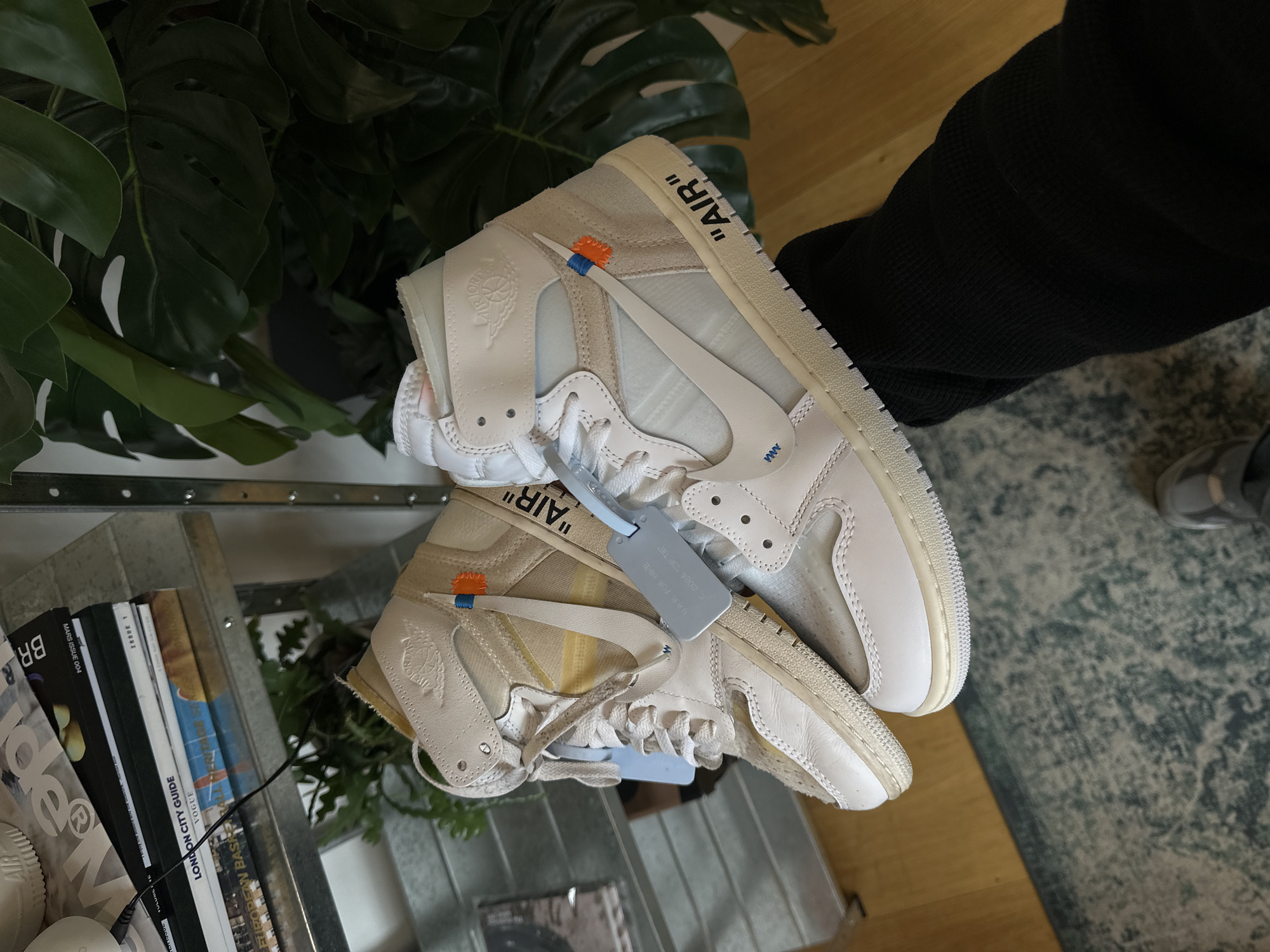The image shows a pair of "Off-White" Air Jordan 1 sneakers, with one shoe stacked on top of the other. The shoes are cream and white with the signature "AIR" branding on the side. There is a tag attached to the bottom shoe. The shoes are being held up with a black sweater in the upper-right corner. To the left is a plant. Below the plant is a metal shelf holding several books. One of the books is titled "LONDON CITY GUIDE".