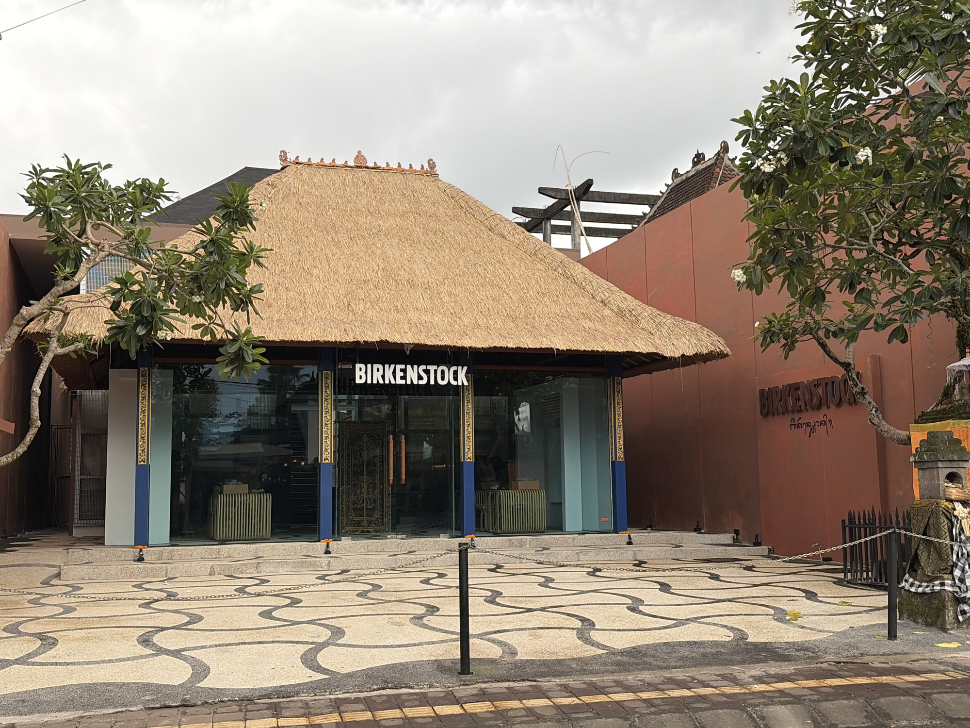 The image shows a Birkenstock store in Ubud District, Indonesia. The store has a traditional Balinese thatched roof and large glass windows. The exterior walls are painted a reddish-brown color. The ground in front of the store is paved with a decorative pattern of interlocking curves. A chain fence separates the store's entrance from the street. A tree with green leaves and white flowers is visible to the left of the store. There are no people visible in the image. The sky is overcast.