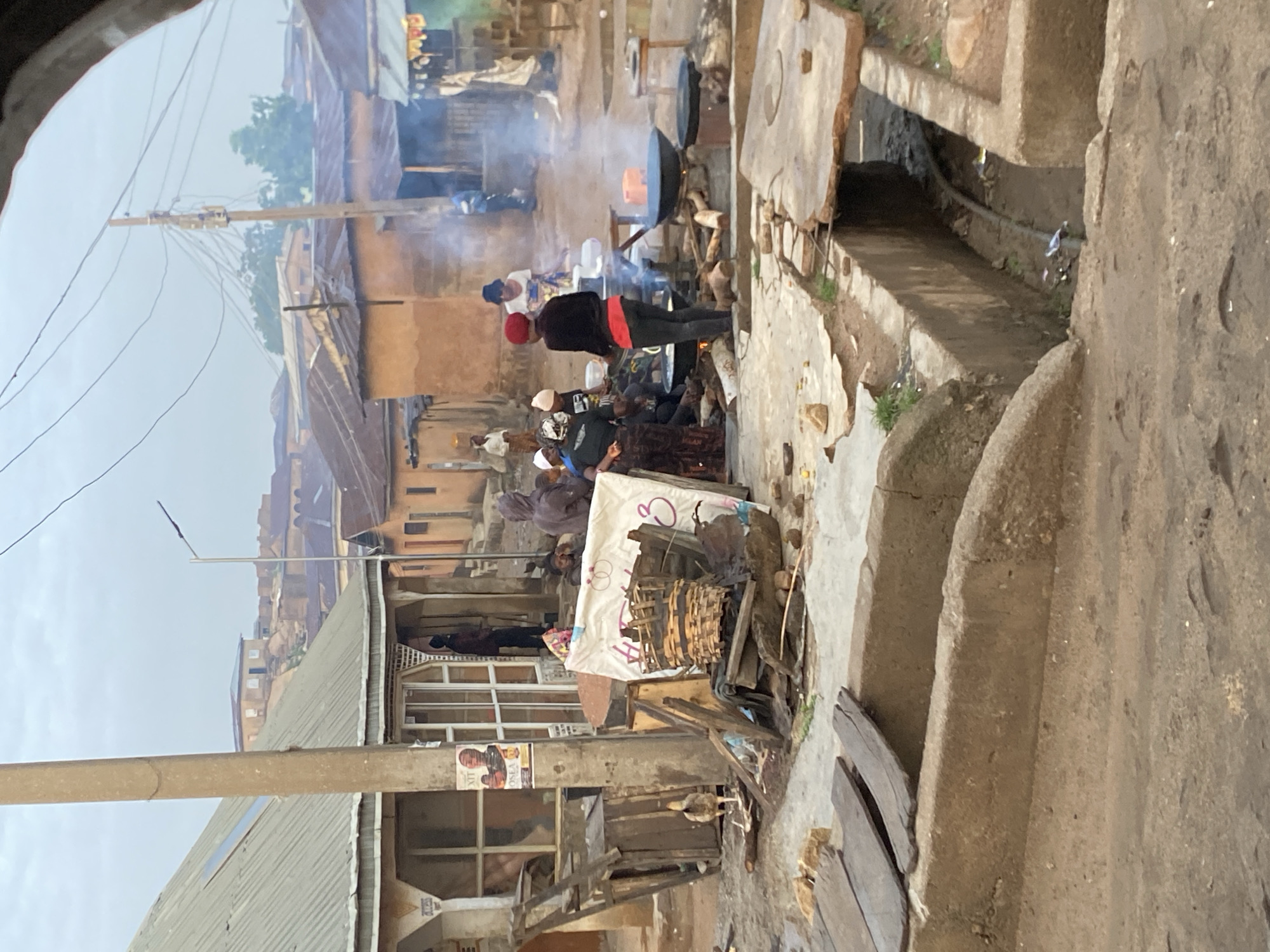 This image depicts a bustling outdoor scene in the city of Gwong, Nigeria, with a group of people engaged in cooking activities. The environment appears to be a residential or market area, characterized by traditional housing with corrugated metal roofs and earthen walls. In the foreground, there is a large open drain or trough, partially covered with wooden planks.
The central focus of the image is a communal cooking setup where several individuals are gathered around open fires with large pots. Smoke billows from the cooking areas, indicating active food preparation. One woman, wearing a red headscarf and black top with red pants, stands prominently facing away from the viewer, seemingly overseeing the cooking. Other people are seated or standing around the cooking area, some holding bowls or utensils.
To the left of the cooking area, a small chicken is pecking at the ground near some wooden structures and discarded items. Further left, a building with a corrugated roof and open windows or verandas is visible, with a poster affixed to a utility pole. In the background, more buildings and a hilly landscape can be seen under an overcast sky, suggesting the general topography of the region. The overall impression is one of a vibrant, everyday community gathering, possibly for a meal or a local event.