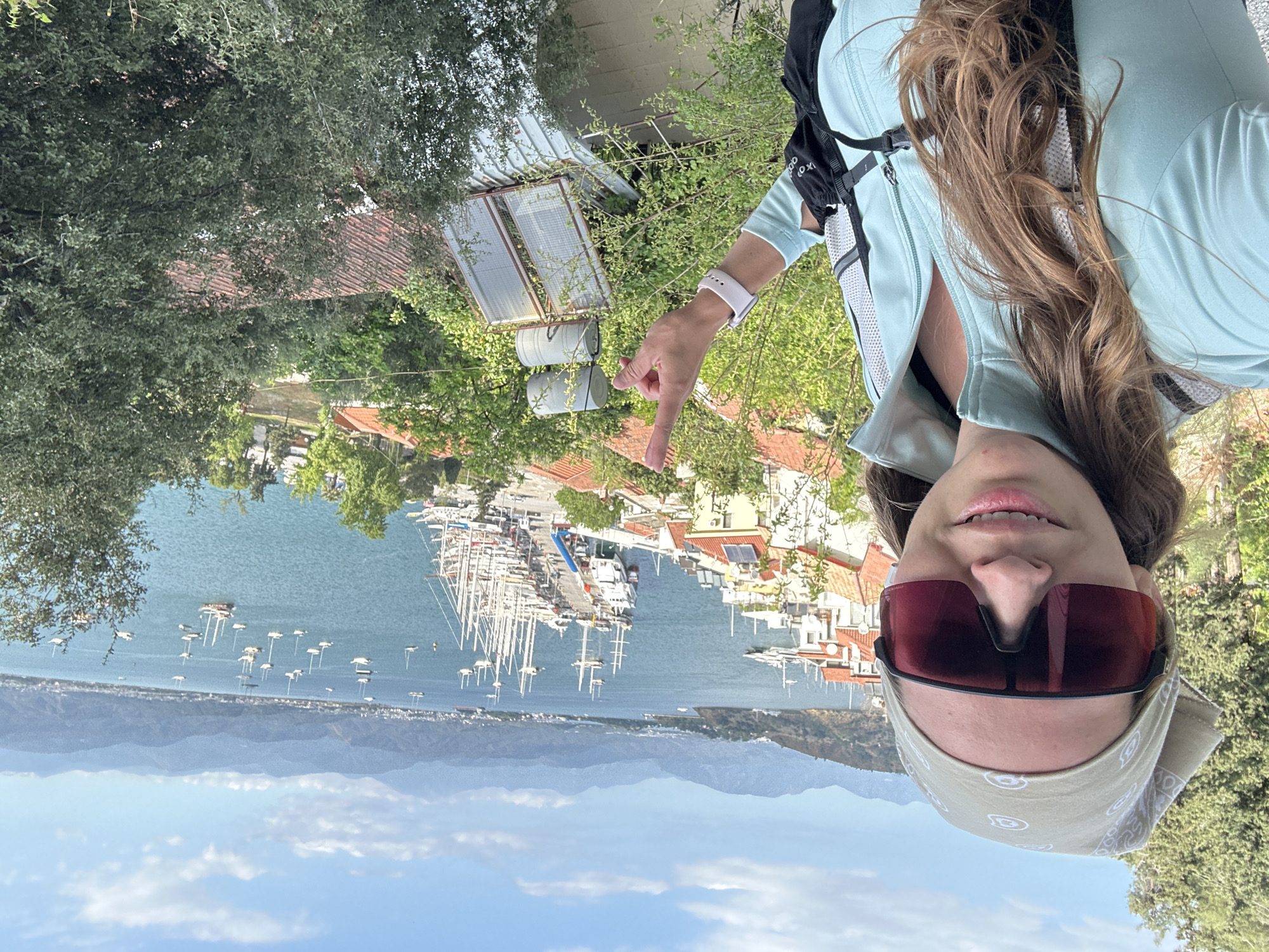A selfie-style, landscape-oriented photograph shows a woman in athletic gear standing on a hill overlooking Fethiye, Turkey. The camera faces directly at the woman, capturing her from the shoulders up. She wears dark red sunglasses and a light-colored bandana. She is dressed in a pale blue athletic jacket and has a backpack on. She points with her right index finger towards the harbor.
In the background, below the woman, is a wide view of the city and its bay. The blue water of the harbor is filled with many sailboats and yachts. The harbor is bordered by buildings with red tile roofs. In the far distance, a range of mountains lines the horizon. A patch of blue sky with some clouds is visible above the mountains. Green foliage and some infrastructure appear on the hill in the immediate foreground.