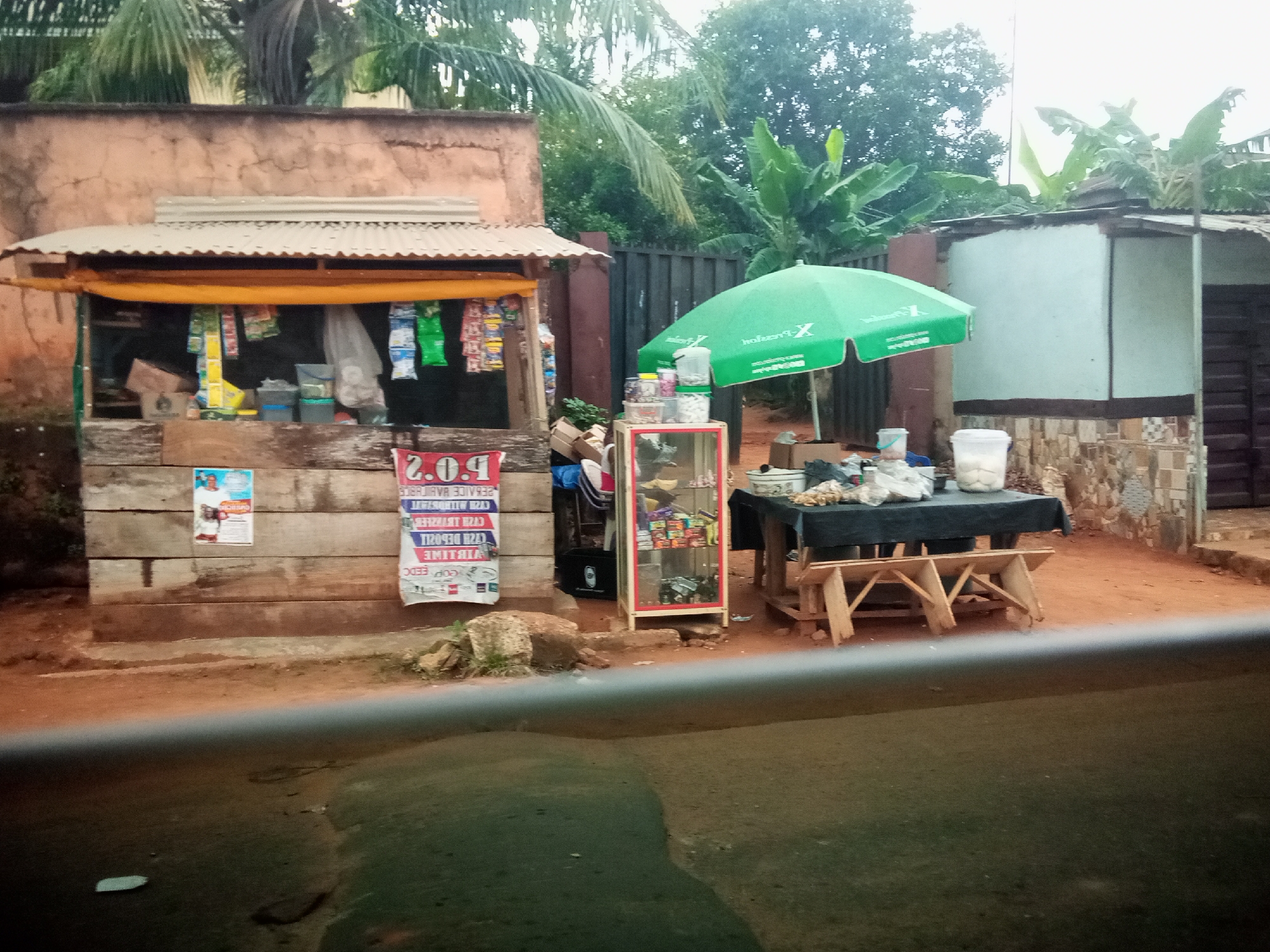 This is a forensic, matter-of-fact description of an outdoor scene, likely a small street-side market, in Nibo, Nigeria.

The scene depicts a weathered, open-air stall with a corrugated metal roof supported by a wooden structure. The stall displays various goods, including bagged items hanging from the roof and items within a small glass display case. Behind the stall, a building with an earthy-colored facade is visible, with lush green trees and foliage in the background.

To the right of the stall, a large, bright green umbrella, branded with white text, is open, providing shade. Beneath the umbrella, a table covered with a dark cloth is set up, displaying more items, possibly food or goods for sale. A wooden bench or seating structure is placed underneath the table. Several white buckets or containers are visible on the ground and on the table.

In the foreground, the ground appears to be a mixture of dirt and possibly a paved surface, with some vegetation growing. The overall impression is of a humble, functional commercial space in a tropical or subtropical environment. No people are clearly visible in the image, nor are any specific actions or interactions taking place. The lighting suggests it is daytime, possibly overcast or late afternoon, given the soft shadows.