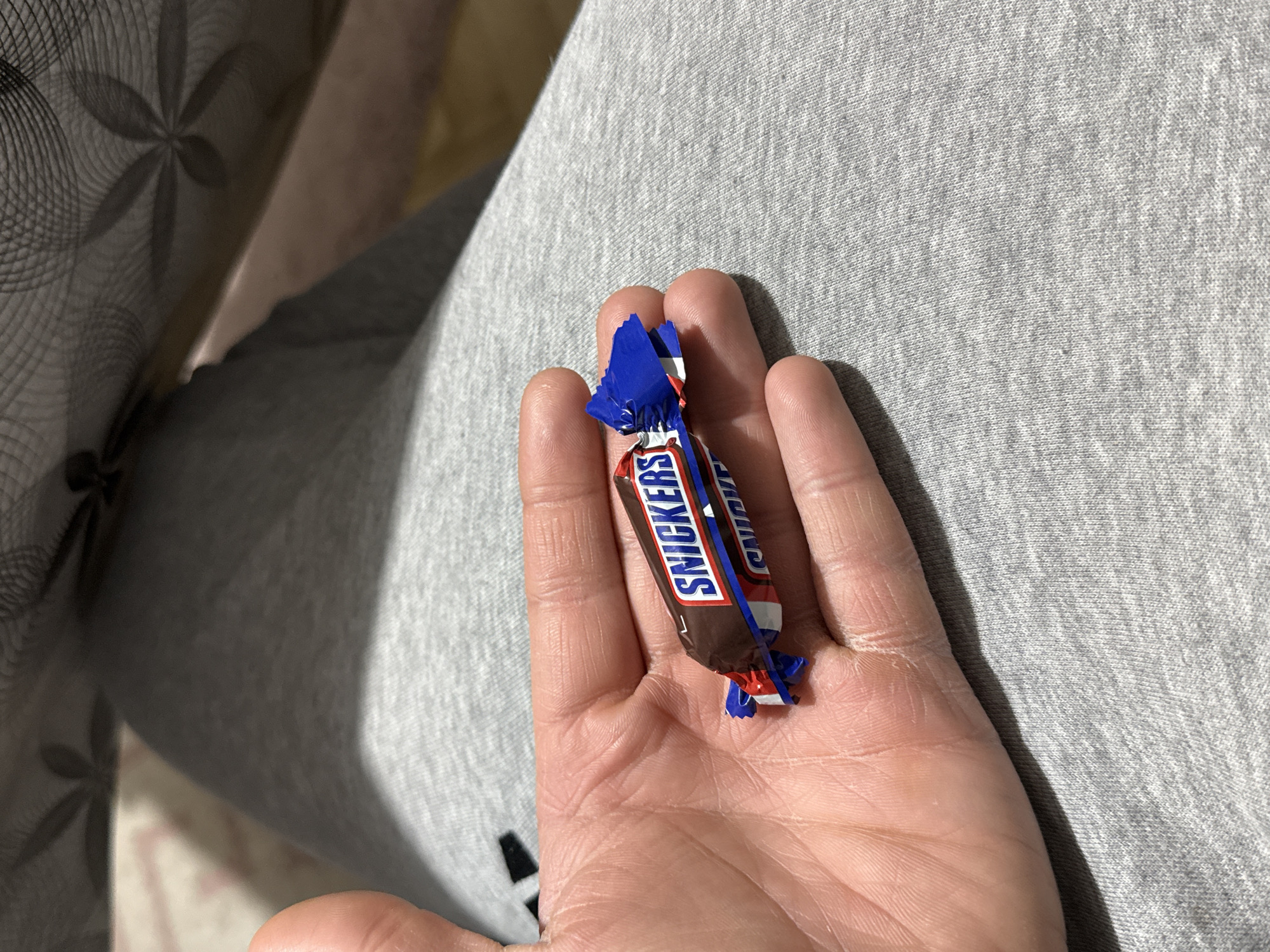 A partially opened Snickers bar rests on the palm of a hand. The hand is resting on a gray, textured surface, possibly clothing. A decorative fabric with a floral pattern is visible to the left. There is no information in the image to determine the location is Stara Zagora, Bulgaria.