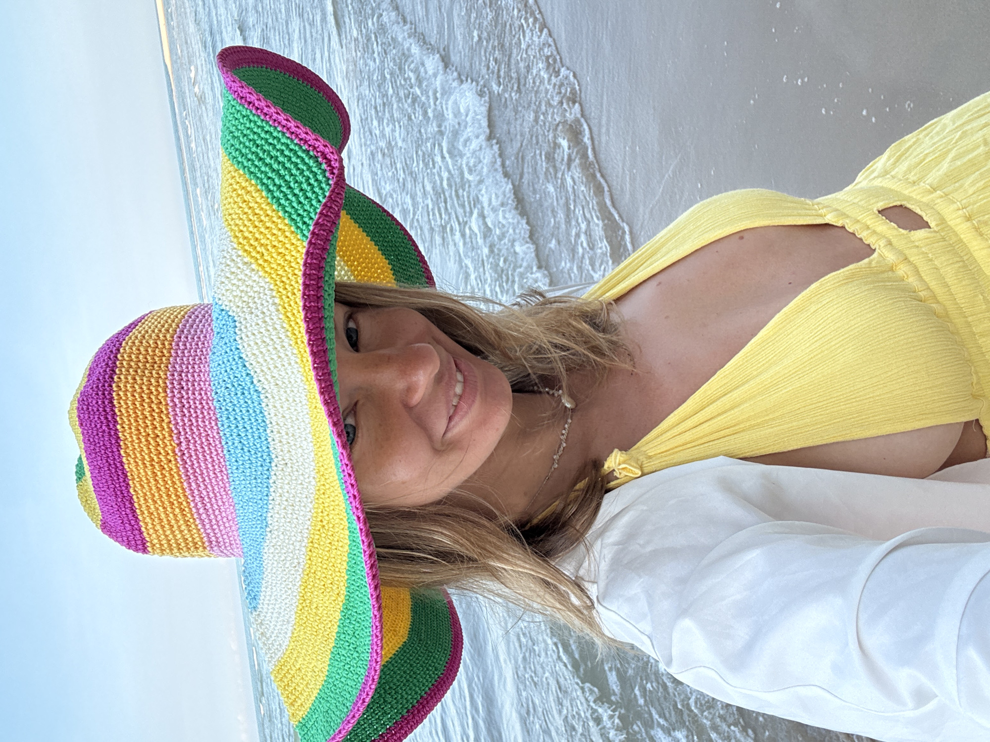 A woman stands on a sandy beach in front of the ocean, wearing a wide-brimmed, multi-colored striped hat, a yellow dress with a deep V-neck, and a white jacket draped over her shoulders. Her blonde hair is visible around the hat, and she is smiling at the camera. The ocean waves are gently rolling onto the shore. The scene is set during daylight, with a clear sky and the sun casting soft light. The location is identified as the city of Thuận Thành, Vietnam.