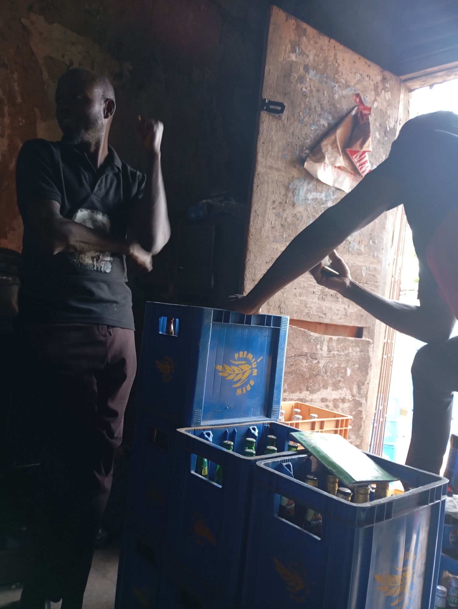 The image shows the interior of what appears to be a bar or storage area in Shendam, Nigeria. Two individuals are visible. The person on the left, a bald man with dark skin and a goatee, is wearing a black polo shirt and dark pants. He is looking to his left with his right arm raised and fist clenched, suggesting an engaged or assertive stance. The person on the right, partially obscured and facing away from the camera, is wearing dark clothing and has their arms extended, holding a mobile phone in their right hand.
In the foreground, there are stacks of blue plastic crates. Some of these crates contain glass bottles, which appear to be beverages. The crates are labeled with yellow emblems and text, some of which are partially visible. Another orange crate is visible behind the blue ones, also containing bottles. The environment is dimly lit with rough, textured walls and a rustic feel. The background shows an open doorway or passage with sunlight filtering through, and some structures that suggest it's part of a building. A crumpled paper bag or wrapping is attached to the wall near the opening. The overall impression is a functional, possibly informal, commercial setting in Shendam.