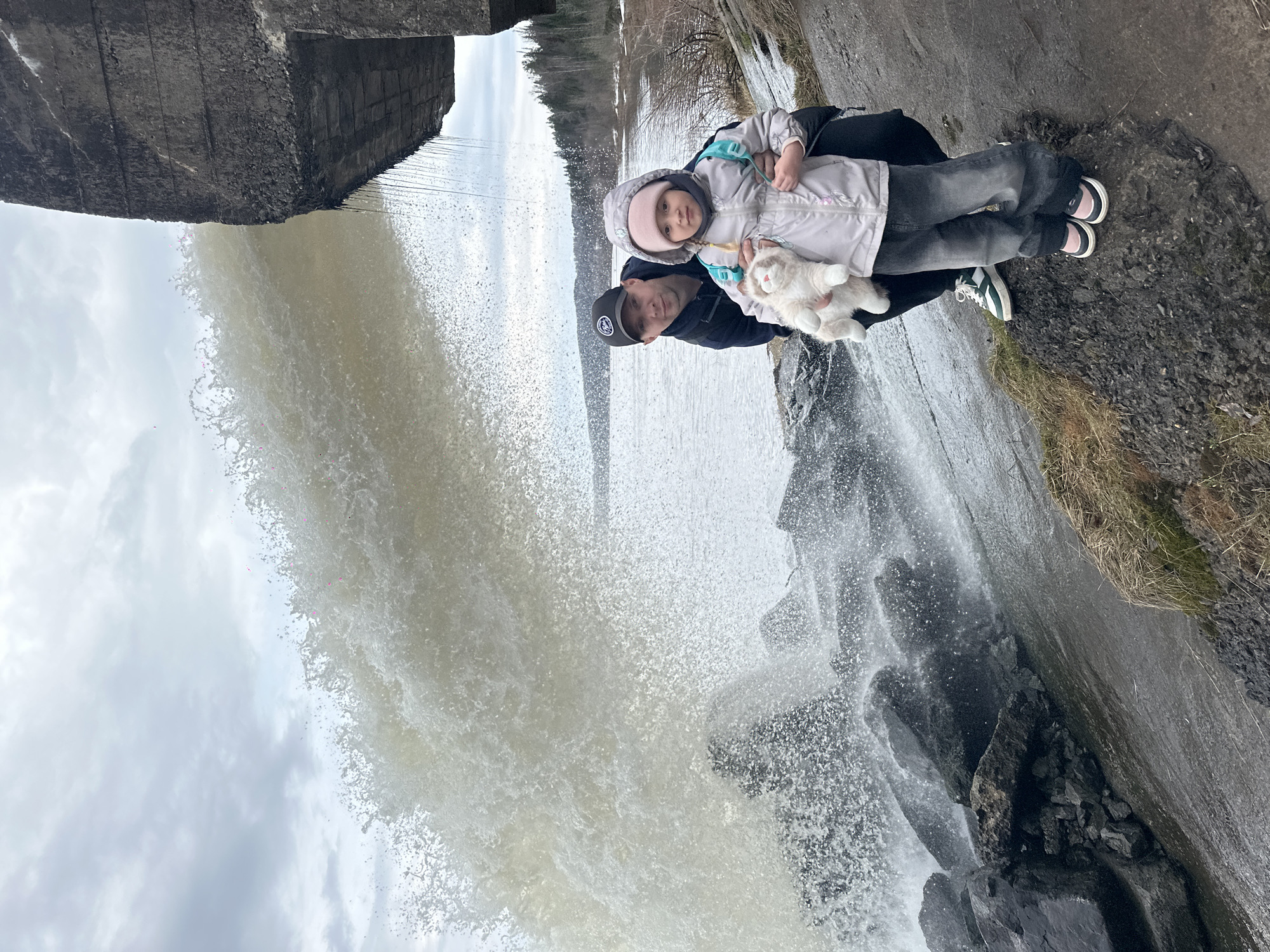 A photograph shows a man holding a child in front of a large waterfall. The man wears a dark jacket and a baseball cap. The child is dressed in a light-colored coat and a hat, holding a stuffed animal. They are positioned on a rocky ledge near the waterfall, with water cascading down in the background. The setting appears to be outdoors, possibly in a natural or park-like environment. The photograph is taken near the Girvas waterfall in Noviy, Russia.