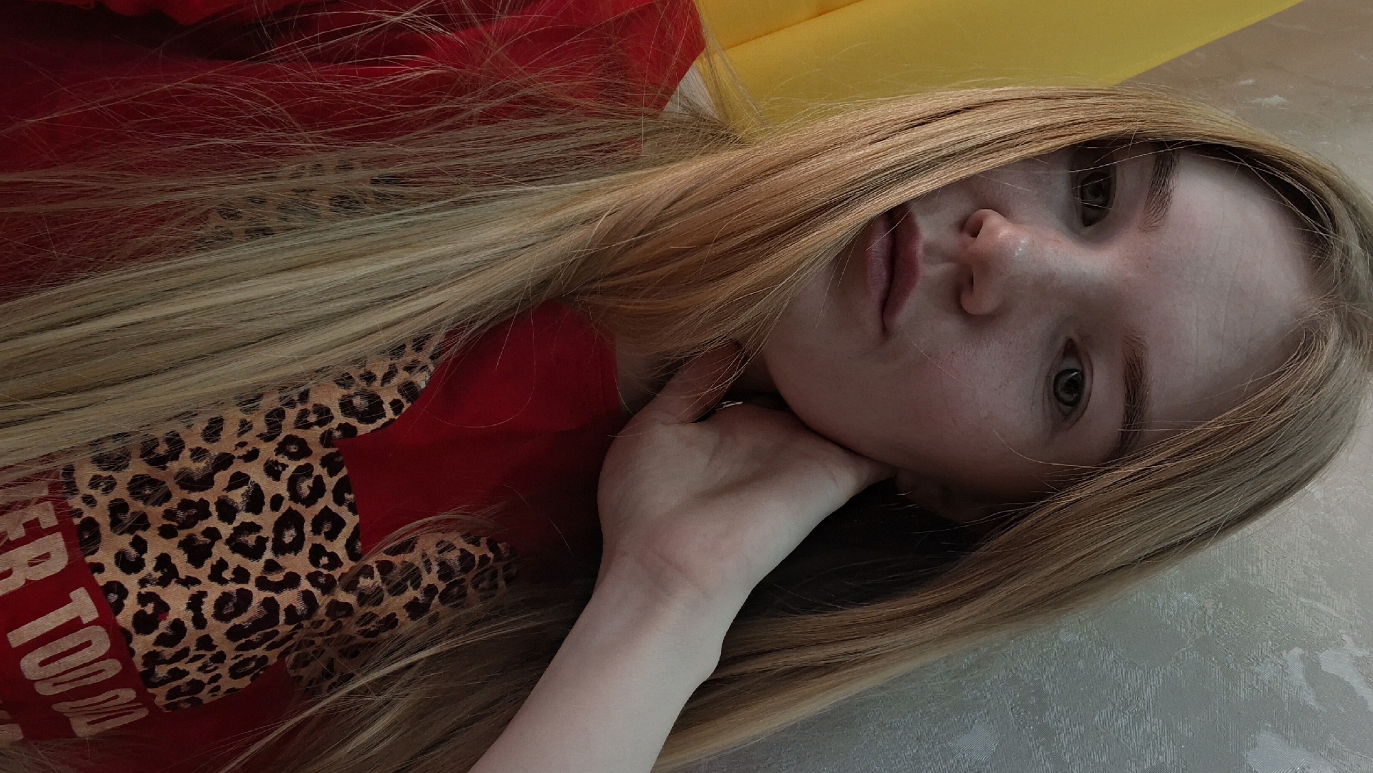 A young woman with long, blonde hair is reclining with her face tilted towards the camera. She rests her face on her hand. She is wearing a red shirt with a leopard-print design. The background includes a yellow surface and what appears to be a textured wall or surface. The location is Chaikovsky, Russia.
