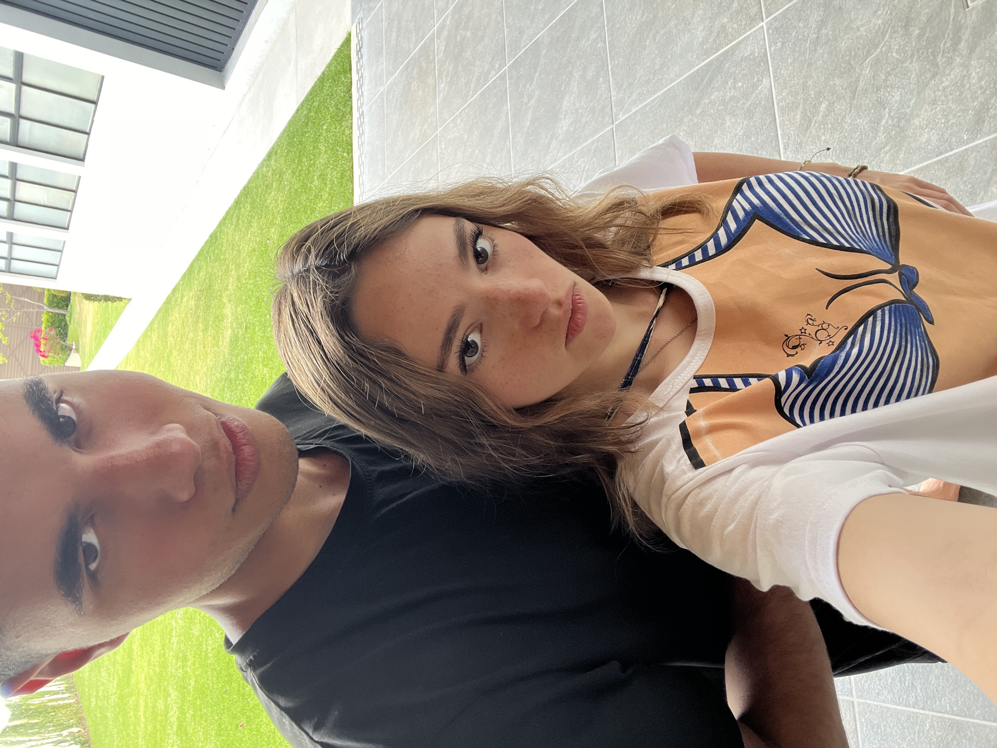 A selfie-style photograph captures two individuals outdoors in San Bernardino Tlaxcalancingo, Mexico. A young woman with shoulder-length light brown hair and fair skin is lying on a man, her face directed at the camera. She is wearing a light orange tank top with a stylized blue and white striped bikini top design printed on it. A silver necklace is visible around her neck. The man has dark hair and skin and is wearing a black t-shirt. The background includes a building with white walls and windows, a well-maintained green lawn, and a gray tiled patio or walkway. The orientation of the image suggests the camera was held at an angle looking down.
