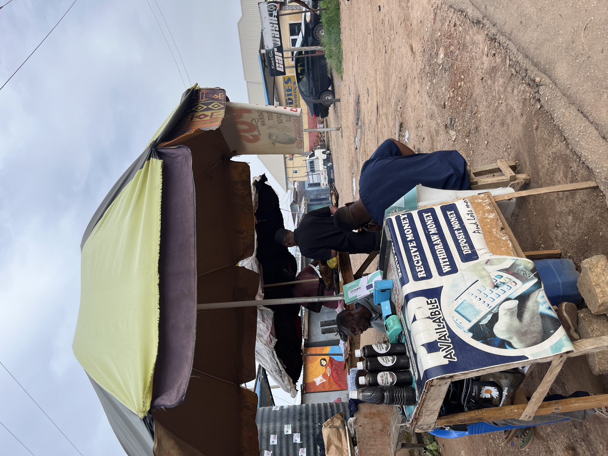 This image captures a street scene in Gura Top, Nigeria, likely during the daytime under an overcast sky. The primary focus is a makeshift stall with a large, colorful tarp providing shade. Underneath, two individuals are engaged in activity. One person, wearing a blue garment, sits near the stall, partially obscured. Another person, in darker clothing, is actively working behind the stall.
The stall itself is laden with various items, including several bottles of a dark liquid labeled "Honey," electronic payment devices (like an "Opay" terminal), and a large blue sign advertising financial services. The sign features text such as "AVAILABLE," "SEND MONEY," "RECEIVE MONEY," "WITHDRAW MONEY," and "DEPOSIT MONEY," indicating that the stall serves as a financial service point. A visual on the sign depicts a hand holding a payment terminal. In the background, a "Car Wash" establishment with cars is visible, suggesting a bustling commercial area. The ground is a mixture of dirt and pavement, typical of many street-side locations in Nigeria. The overall impression is one of everyday commerce and community services operating in an open-air market setting.
