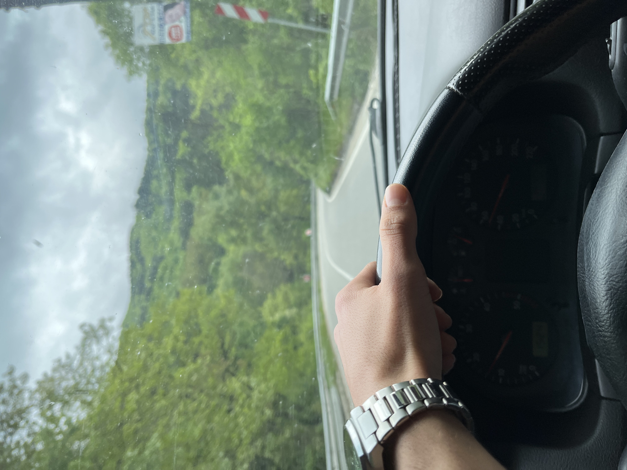 The image depicts a person driving a car, as seen from the driver's perspective. The person's hand is visible, holding the steering wheel. A silver watch is on their wrist. The dashboard is partially visible, showing gauges. Through the windshield, a view of a winding road through a green, mountainous or forested area is seen. A sign with a red and white striped pattern is visible. Based on the image, it is not possible to pinpoint the specific location in Asenovgrad, Bulgaria, only that the car is likely driving along a road with mountainous scenery.