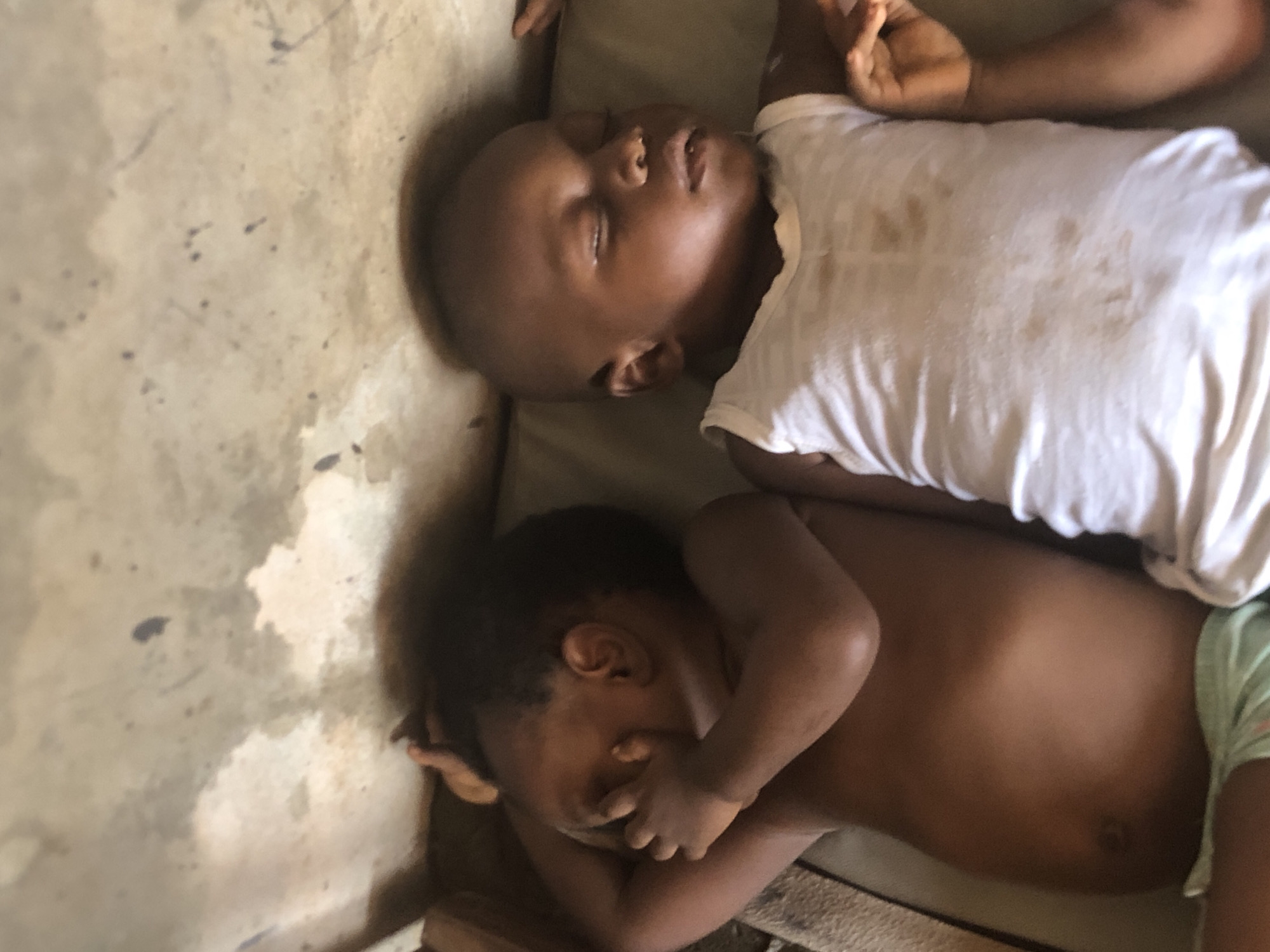 Two young Black children, likely toddlers, are asleep on a cot in what appears to be a modest dwelling. The child on the left is lying on their side, with their right arm bent and their hand covering their face. They are unclothed from the waist up, wearing light green patterned shorts. The child on the right is lying on their back, wearing a dirty white tank top. Their eyes are closed, and their mouth is slightly open. The cot appears to be placed on the floor of a room with concrete walls and a ceiling that shows signs of water damage and staining. The overall lighting suggests natural light entering the space. The setting is likely within Ukwu-Abwa, Nigeria, given the context provided.