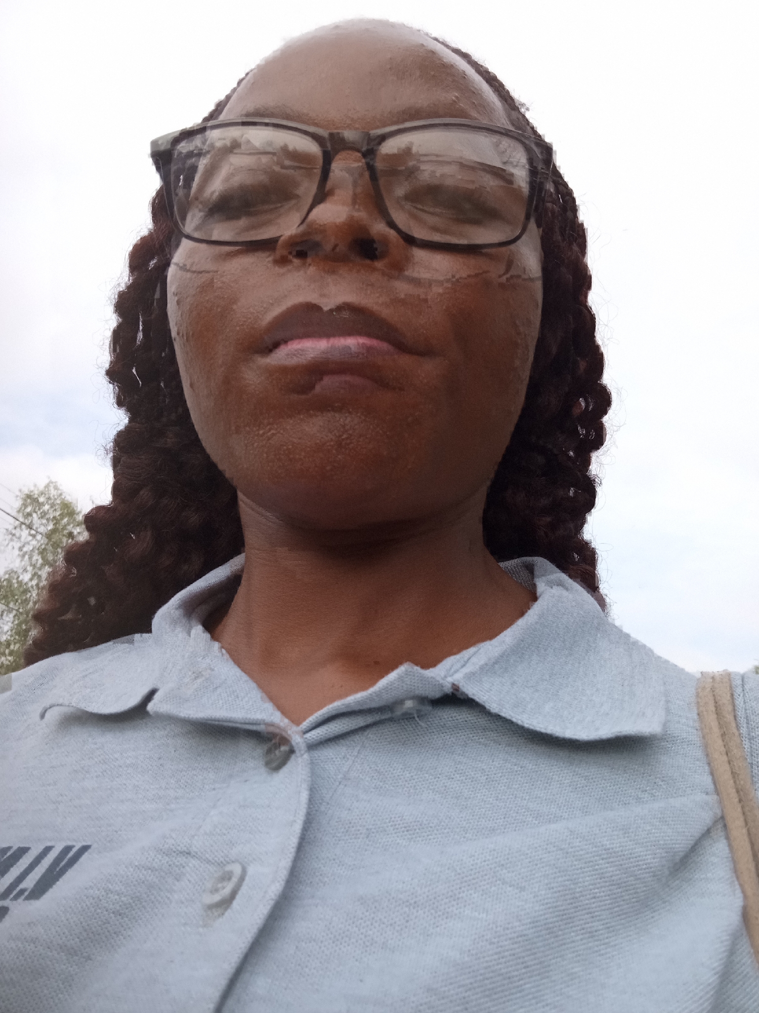 The image shows a close-up, low-angle selfie of a dark-skinned woman wearing glasses and a light blue collared shirt. Her braided hair is visible on the sides of her head. The woman's face is wet, possibly from rain, with water droplets visible on her skin and glasses. There are some darker markings or scars on her cheeks. Her lips appear slightly glossy and pink. The background is a bright, overcast sky with some greenery and power lines visible to the left, suggesting an outdoor setting in Nigeria. The shirt has two light-colored buttons on the placket and a logo or text printed on the left chest area, though the text is partially obscured and unreadable. The overall impression is a candid, informal portrait taken outdoors.