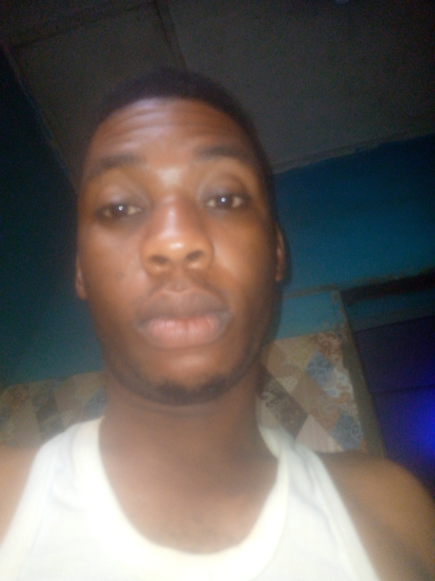 A selfie of a man with dark skin, wearing a white tank top. The background includes a blue wall and a patterned object that could be a pillow or blanket. Location: Unknown, Unknown.