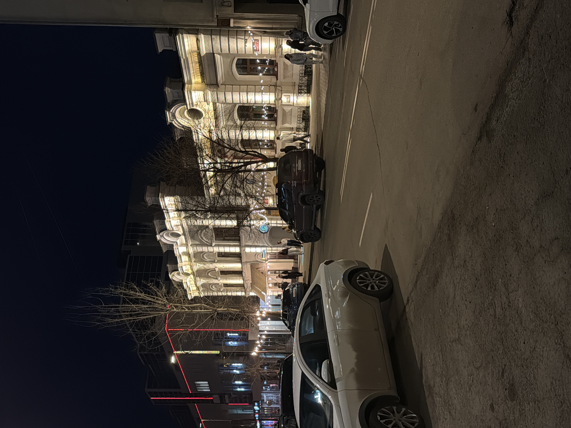 The image captures a vibrant night scene on a city street, likely in Krasnoyarsk, Russia, looking towards a row of buildings. A large, ornate, multi-story building with classical architectural elements, featuring arches, columns, and decorative moldings, dominates the right side of the frame. This building is brightly illuminated with warm artificial lights, highlighting its details and suggesting it houses businesses or residences. String lights are strung across the street or between structures, adding to the festive evening atmosphere.
On the left, another building is visible, featuring modern glass sections and distinct red neon lighting, creating a contrast with the older architecture. Bare tree branches, indicative of winter or early spring, are interspersed between the street and buildings.
Several cars are parked along the street. In the immediate foreground, a white sedan is parked on the right side of the road, partially obscuring the view. Behind it, a dark SUV or car is also parked. The asphalt road shows white double-line markings.
Multiple groups of people are visible on the sidewalk in front of the illuminated building, some standing and conversing, others appearing to walk. Their presence, along with the bright lighting and decorative string lights, suggests an active urban evening environment. The dark sky confirms it is nighttime. No specific text on signs is clearly legible, though a Russian-style license plate can be seen on the dark SUV, its characters are unreadable.