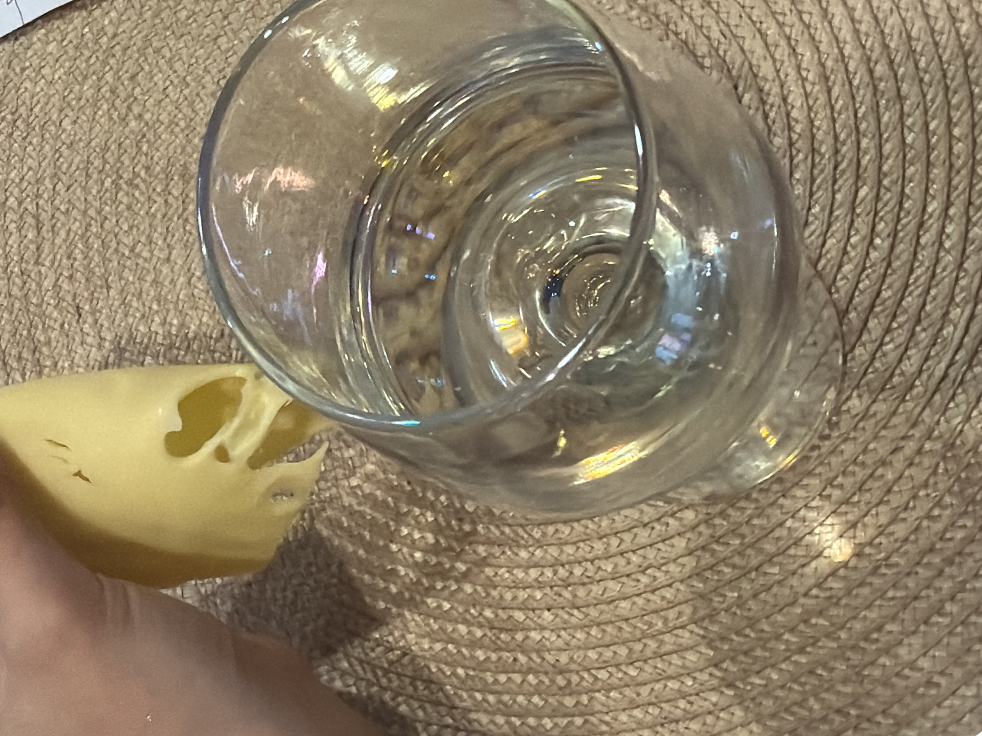 The image depicts a clear glass of water positioned on a woven, possibly straw, placemat or surface. A hand holds a piece of yellow cheese with holes near the glass. No elements in the image connect it to the city of Pervomaysky, Russia.