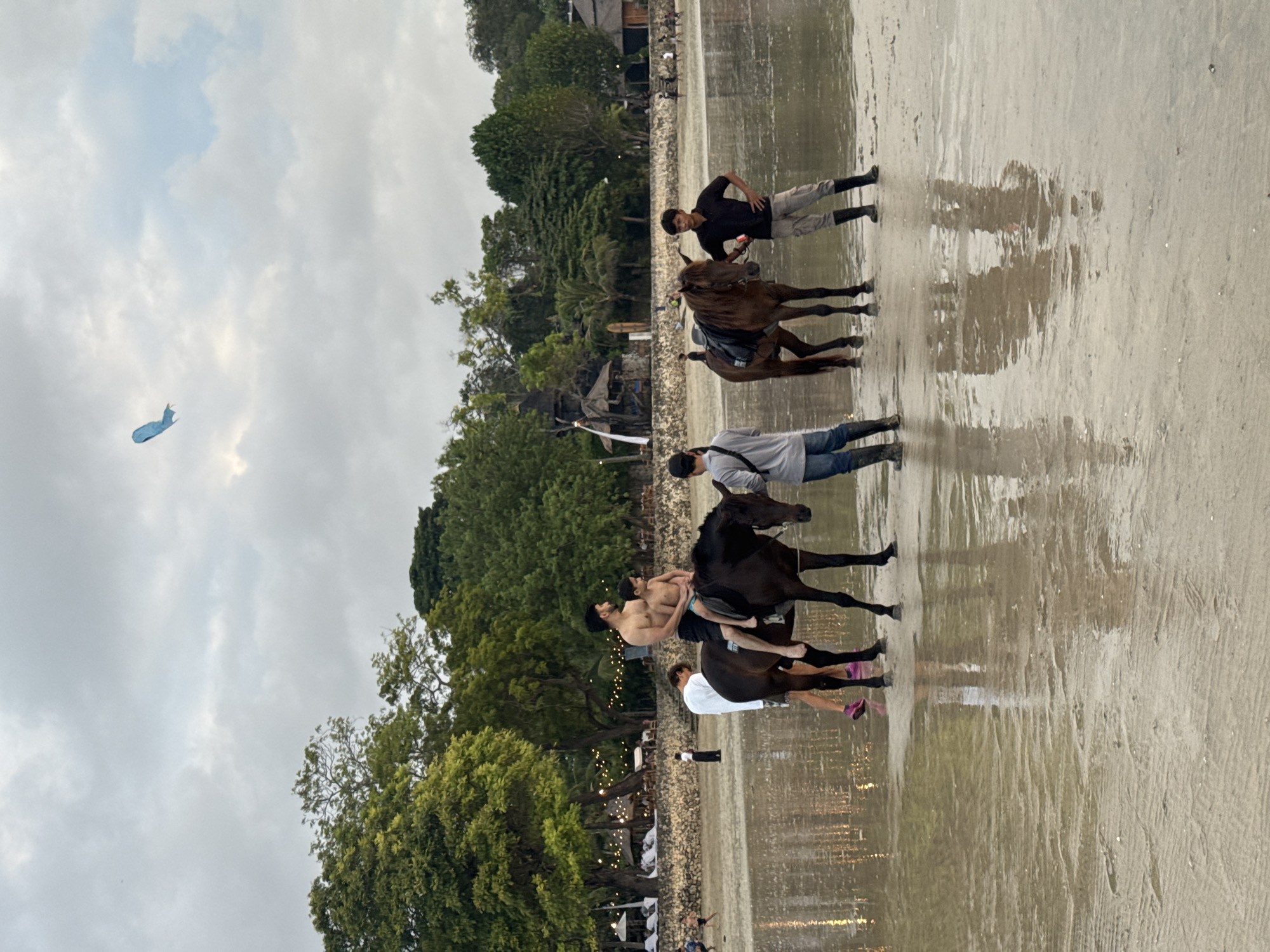 In Kuta Selatan, Indonesia, on a wet sandy beach, two brown horses stand in shallow water. On the dark brown horse to the left, a shirtless man with dark hair sits in front, and a young boy with short dark hair sits behind him. The man is wearing dark shorts, and the boy is wearing blue shorts. Another man, wearing a white long-sleeved shirt and dark shorts, stands to the left of this horse, facing away from the camera. To the right of the horses, a man wearing a black short-sleeved shirt and light-colored pants, with dark boots, stands next to the second horse, holding its reins. Another man, wearing a gray long-sleeved shirt and blue jeans, stands in front of the second horse, facing away from the camera. The horses are equipped with saddles. In the background, lush green trees and various structures, possibly resorts or restaurants, are visible. The sky is cloudy, and a light blue kite is visible in the upper portion of the frame.