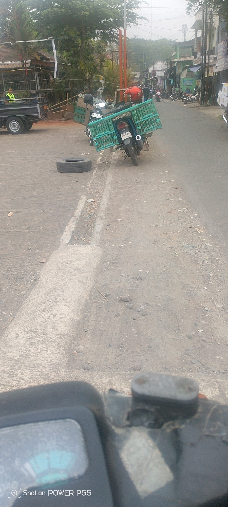 An eye-level, medium shot captures a dirt road in an urban environment, with a focus on a motorcycle carrying a large green crate. The motorcycle is parked facing right, with its rear wheel visible. The green crate appears to be made of plastic and is loaded with what seems to be goods. A spare tire is lying on the ground next to the motorcycle. In the background, a black truck is visible on the left, and further down the road, several motorcycles and a few people are present, suggesting a busy street. The road is unpaved, with scattered stones and debris. The sky is overcast, and the overall lighting is dim. The foreground is dominated by a portion of a motorcycle's dashboard and handlebars, indicating the photographer is on another motorcycle.