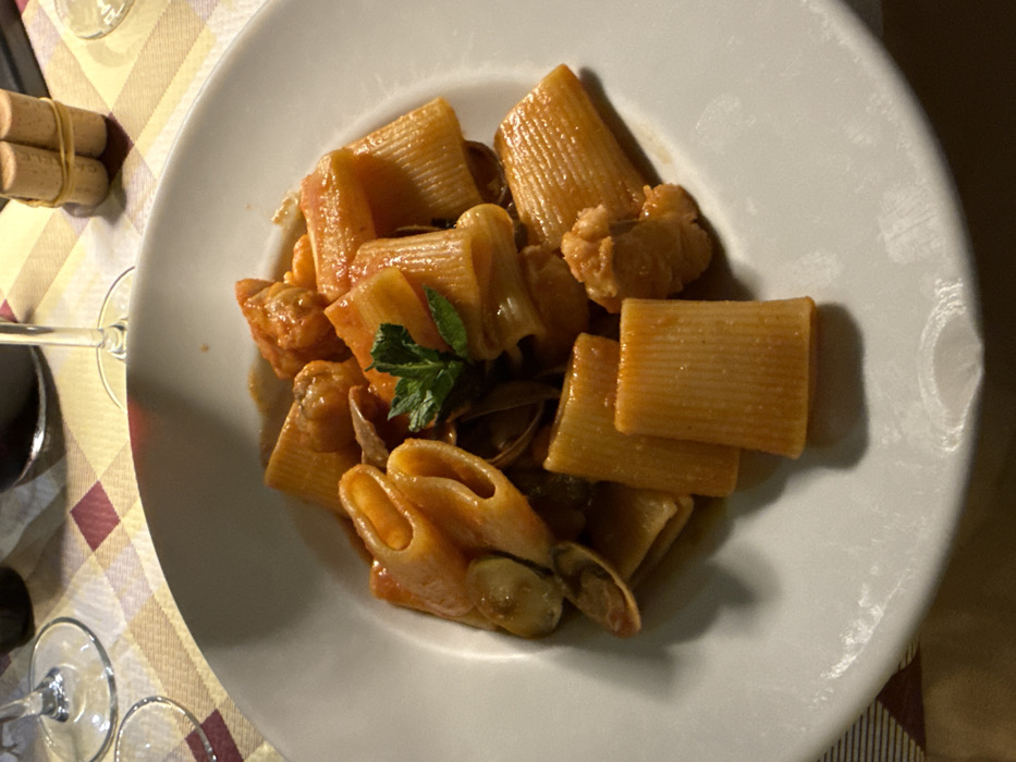 A close-up, elevated view captures a white ceramic plate containing a serving of rigatoni pasta, coated in an orange-red seafood sauce. The dish includes whole clams, some open, and irregular pieces of white seafood, likely monkfish. A small sprig of fresh mint garnishes the center. The plate rests on a checkered tablecloth featuring squares of light brown, white, and maroon. Partially visible in the background are two clear wine glasses, one containing red wine, and a small bundle of wine corks secured with cord, indicating a dining setting within a restaurant in the city of Lecce, Italy. No individuals are visible.