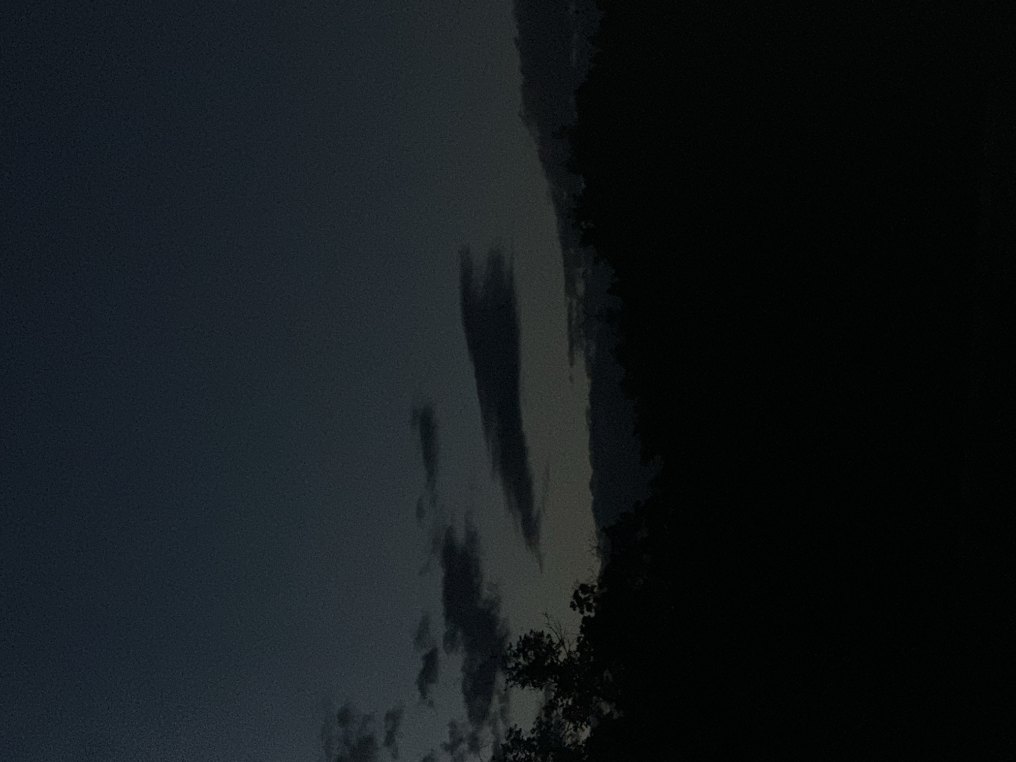 The image depicts a dark, atmospheric scene with a focus on the sky. The time of day appears to be dusk or early night, given the dim lighting and deep shadows. The sky is a muted, dark blue-gray, with scattered clouds that are also dark and elongated, suggesting movement or a particular cloud formation. A dense, dark mass on the right side of the image likely represents trees or a forest, creating a stark silhouette against the slightly lighter sky. The overall mood is somber and perhaps a bit mysterious, with the limited light and dark contrasts. There are no visible people, objects, or text in the image, the scene is solely of a natural, outdoor environment at twilight. The location context of Nizhny Novgorod, Russia, does not offer specific visual cues in this particular image beyond a general impression of a temperate climate. The weather seems to be clear or partly cloudy, without any indication of rain or strong winds, though the dark clouds could imply impending weather changes.