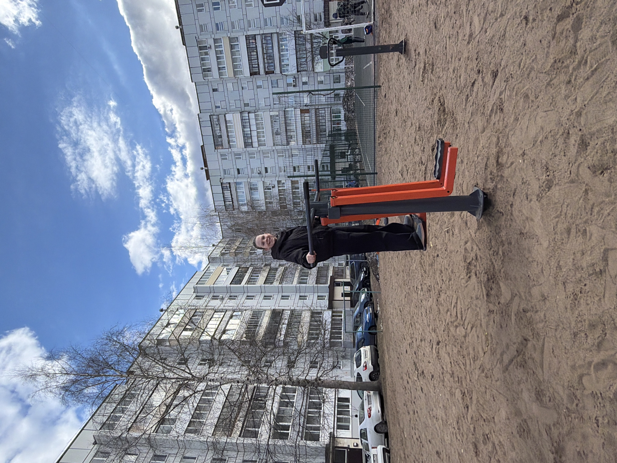 An outdoor, daytime scene shows a young man actively engaged with public exercise equipment in what appears to be a residential courtyard or recreational area. The image is originally rotated, but when viewed upright, the man is positioned towards the center-left, facing slightly towards the viewer, with a neutral to slightly smiling expression. He is dressed in dark clothing, including a black jacket and pants, and is holding onto the handles of an orange and black outdoor gym machine, likely designed for abdominal or leg exercises.
The ground around the equipment is unpaved, consisting of sand or dirt, showing some footprints and textured surfaces. In the background, numerous multi-story residential apartment buildings, typical of Soviet-era architecture, dominate the skyline. These buildings are light-colored, featuring many windows and balconies. Bare trees, indicating either late winter or early spring, are scattered throughout the scene, especially between the buildings and the foreground. Several cars of various colors are parked along what appears to be a driveway or parking area adjacent to the exercise space and buildings.
The sky is bright blue with scattered white, cumulus clouds, suggesting clear and sunny weather. Bright sunlight illuminates the scene, creating some visible shadows. To the right of the man and beyond the exercise equipment, a fence, possibly enclosing a sports court, is partially visible, with hints of a goal post in the distance. The overall impression is one of urban outdoor activity on a bright, cool day. No discernible text is visible in the image. The setting aligns with typical urban environments in places like Chaikovsky, Russia.