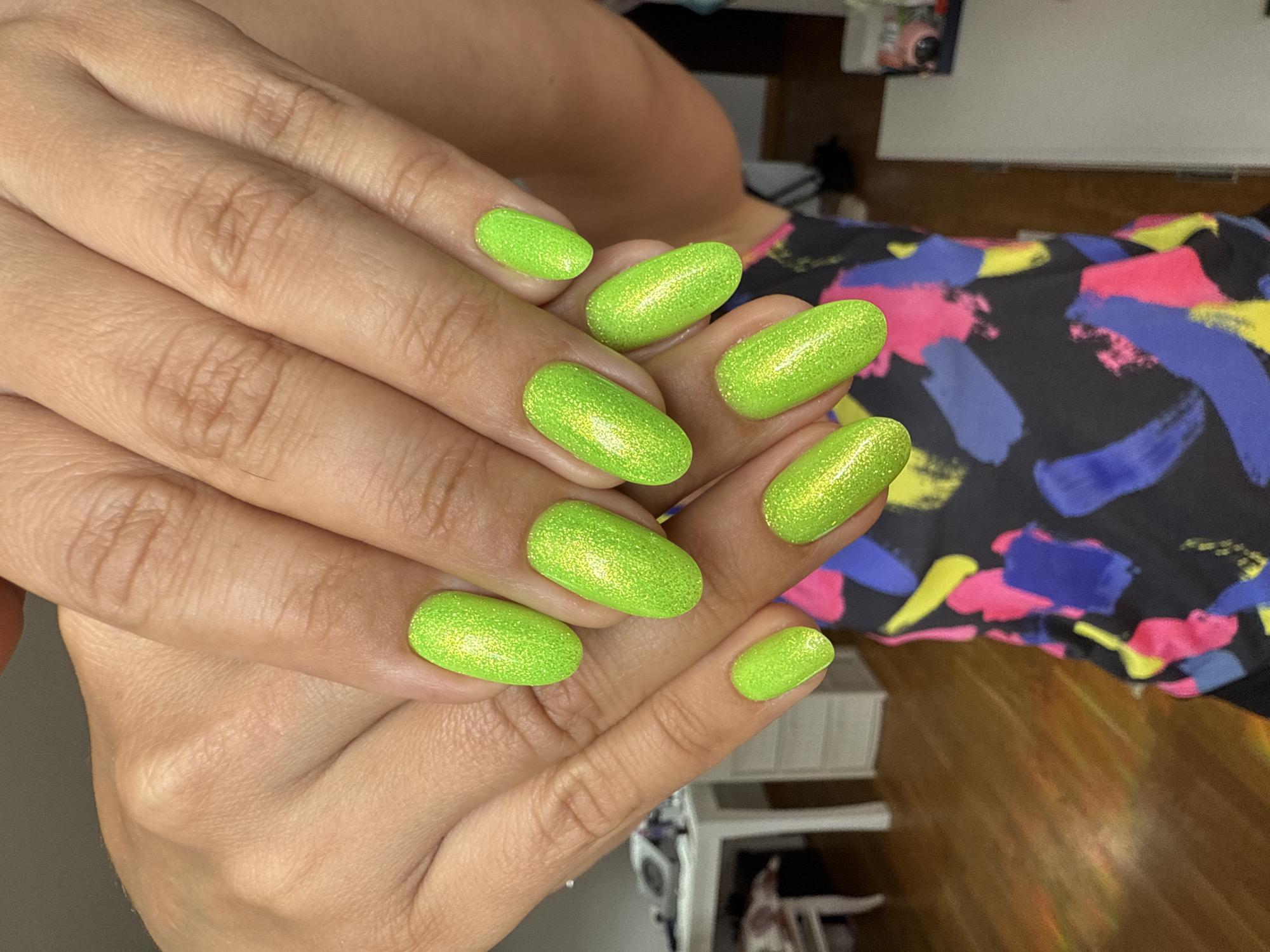 The image shows a pair of hands with bright, neon green, glittery manicured nails. The hands are resting on a patterned black top. In the background, a wooden floor and a white table with a drawer unit are visible. The scene is indoors, likely in a domestic setting. The lighting suggests it might be daytime.