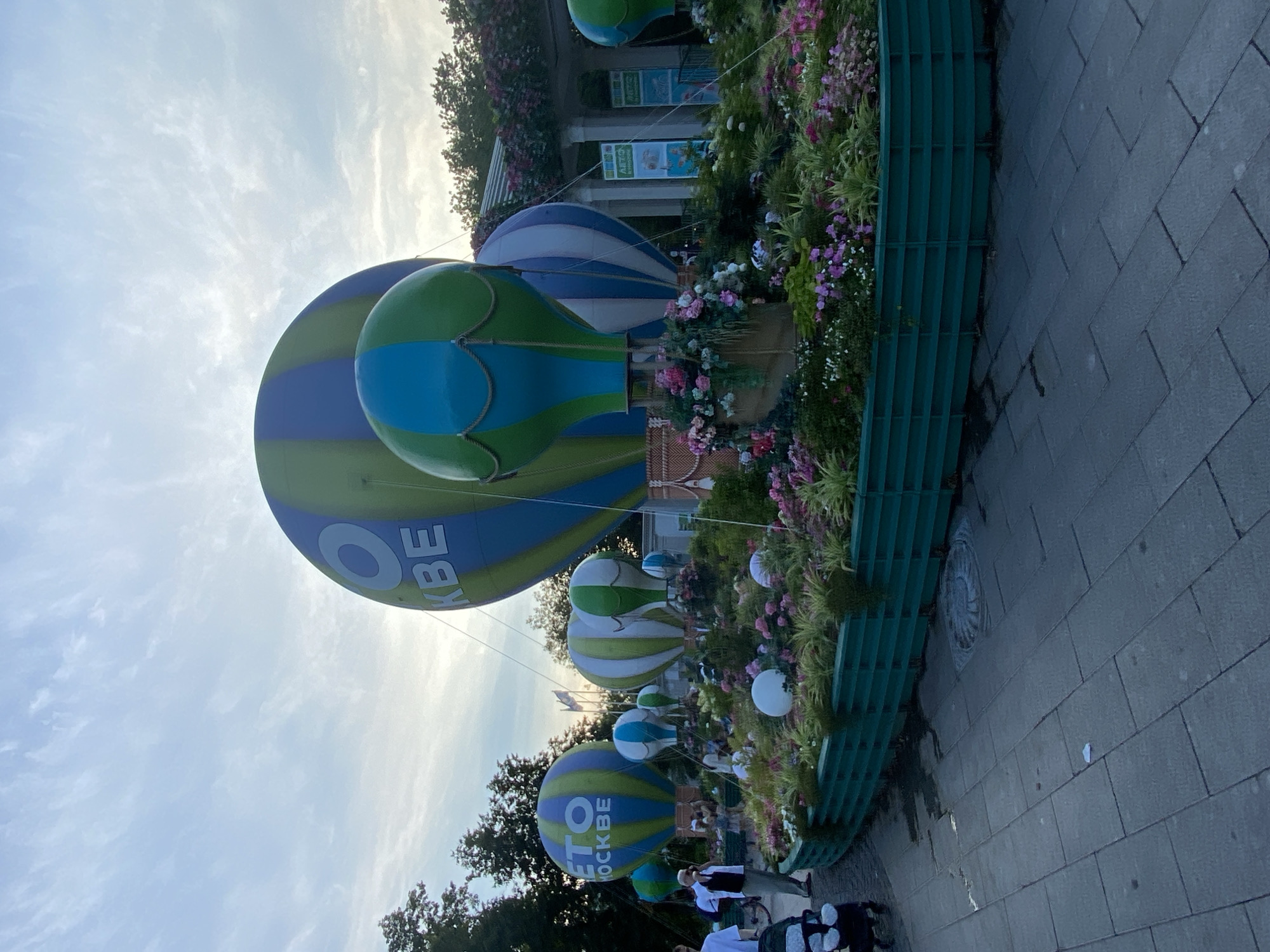 The image shows a festive outdoor display featuring large, colorful hot air balloons and a floral arrangement in a city park. The hot air balloons are primarily green and blue, with some featuring white stripes. One large balloon in the center has the text "О КВЕ" in white letters. Another balloon on the left has the text "ЭТО" and "МОСКВЕ" in white letters. These suggest the display is related to Moscow, Russia. The floral arrangement is vibrant, with pink, white, and purple flowers, as well as lush green foliage. It is contained within a green, segmented barrier. Several people are visible in the background, some walking and others standing. One person appears to be pushing a stroller. The sky is partly cloudy, with the sun low on the horizon, indicating it is likely evening or early morning. The ground is paved with grey rectangular tiles, and there are some trees and buildings visible in the distance. The overall impression is of a public space decorated for an event.