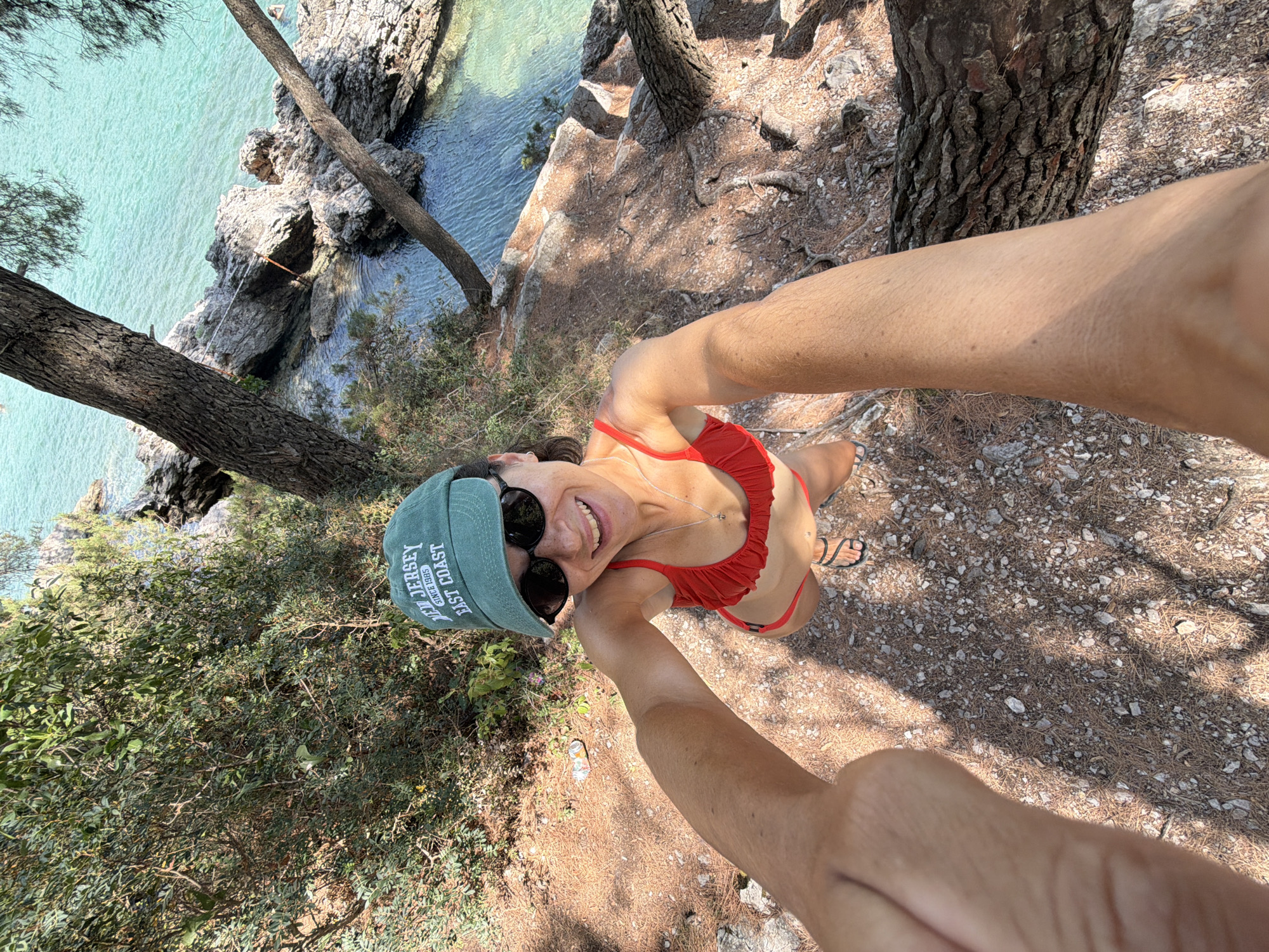 A woman in a red bikini and a green baseball cap is taking a selfie on a rocky, pine-covered cliff overlooking the turquoise waters of the Adriatic Sea in Bigova, Montenegro. The woman is smiling and looking up at the camera, with her arms extended. The sea is clear and calm, with rocky outcrops and small waves lapping at the shore. Trees and foliage surround the cliff, providing shade and a natural setting. The overall impression is one of a sunny day and a relaxed vacation atmosphere.