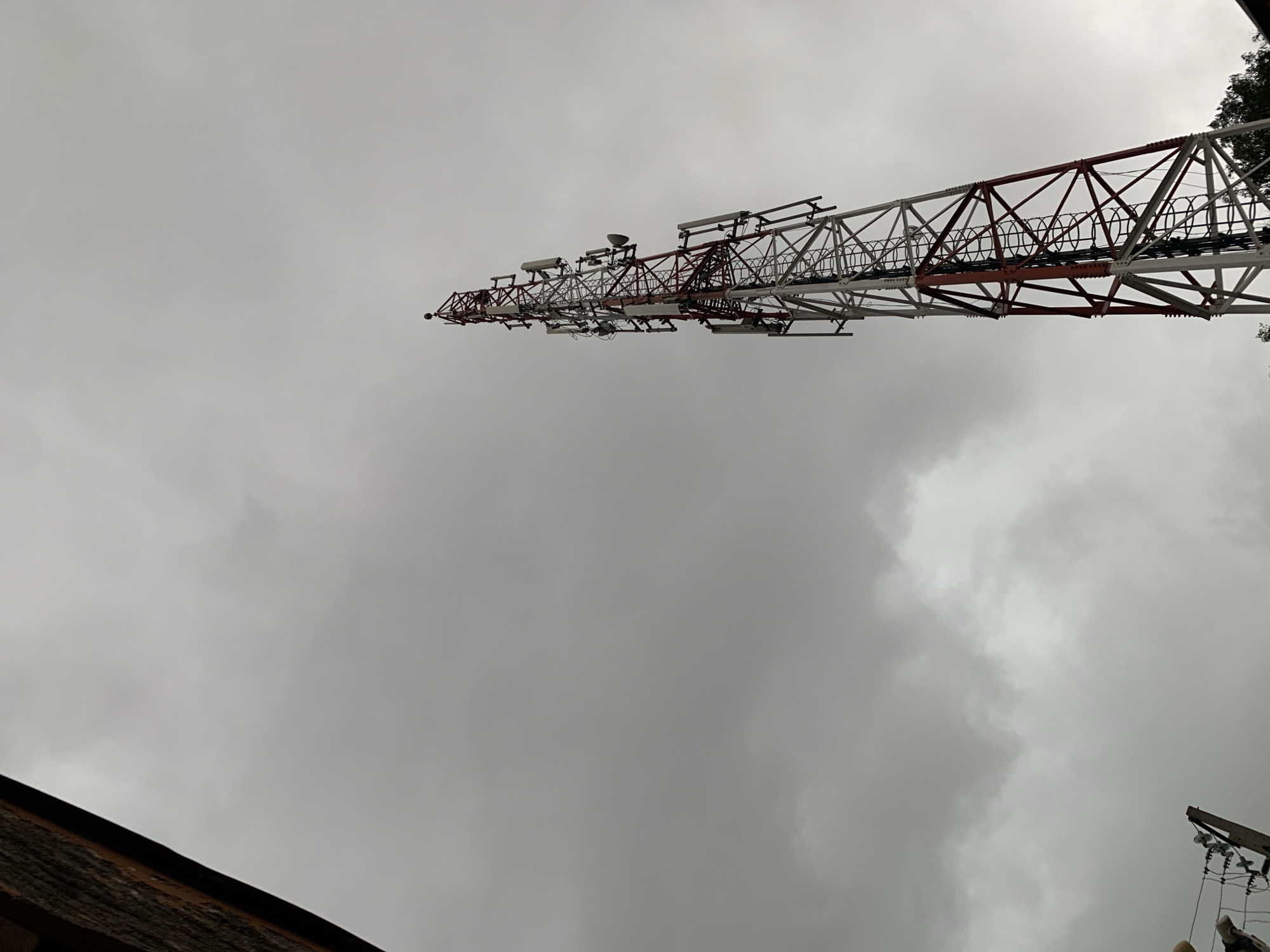 The image displays a large, horizontally oriented metal truss structure, likely a communication or transmission tower, set against a predominantly overcast sky. The structure is composed of an alternating pattern of red (or reddish-brown) and white (or light gray) lattice beams. Numerous antennas and communication equipment, including disc-shaped and rectangular panel antennas, are mounted along its length, especially towards the left side which appears to be its narrower end. Various wires and cables run along the tower.
The sky is almost entirely filled with dense, grey clouds, indicating an overcast day with diffused lighting. In the bottom-left corner, a portion of a brown, possibly tiled or shingled, roof is visible. To the bottom-right, the top section of a wooden utility pole with insulators and electrical wiring is partially captured.
The photograph is taken from a low-angle perspective, looking up towards the sky and the structure. No people are visible, and no specific activity is occurring. The weather is cloudy, suggesting a dull day. There is no discernible text within the image. The scene is located in Rafin Par, Nigeria.