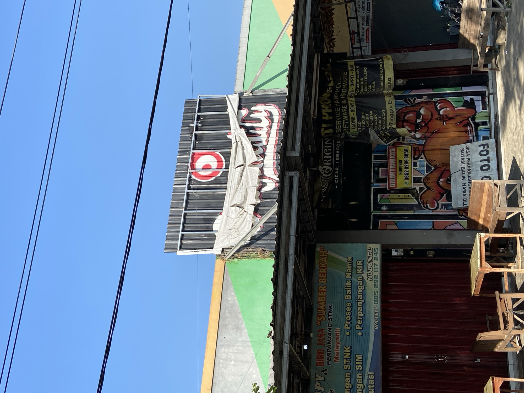 This image depicts a street-level view of businesses in Margorejo, Indonesia. The foreground shows outdoor seating with wooden chairs and tables, suggestive of a cafe or eatery. Behind the seating area, several storefronts are visible. One prominent sign, partially obscured by a torn tarp, displays the logo of a red circle with a white target inside, followed by stylized lettering that reads "JUJU."
To the left, a blue and green banner advertises "OPY & BIRO JASA SUMBER BERKAH," listing services such as "PERPANJANG STNK," "Proses Balik Nama," and "Perpanjangan KIR." Below this, a red roller door suggests an entrance.
To the right of the "JUJU" sign, another storefront features a black banner for "ORIGINTI PHONE," which appears to be a phone repair and sales shop, offering services like "SERVICE HANPHONE," "SOFTWARE & HARDWARE," and listing various phone brands. Adjacent to this is a smaller yellow sign with a phone number, reading "DIKONTRAKKAN HUBUNGI 0852 8289 8058."
Further to the right, a vibrantly painted mural, featuring a character resembling Garfield, covers a significant portion of a shop's facade. The overall impression is of a bustling commercial area with a mix of services and local businesses. The sky above is clear blue with power lines strung across it.