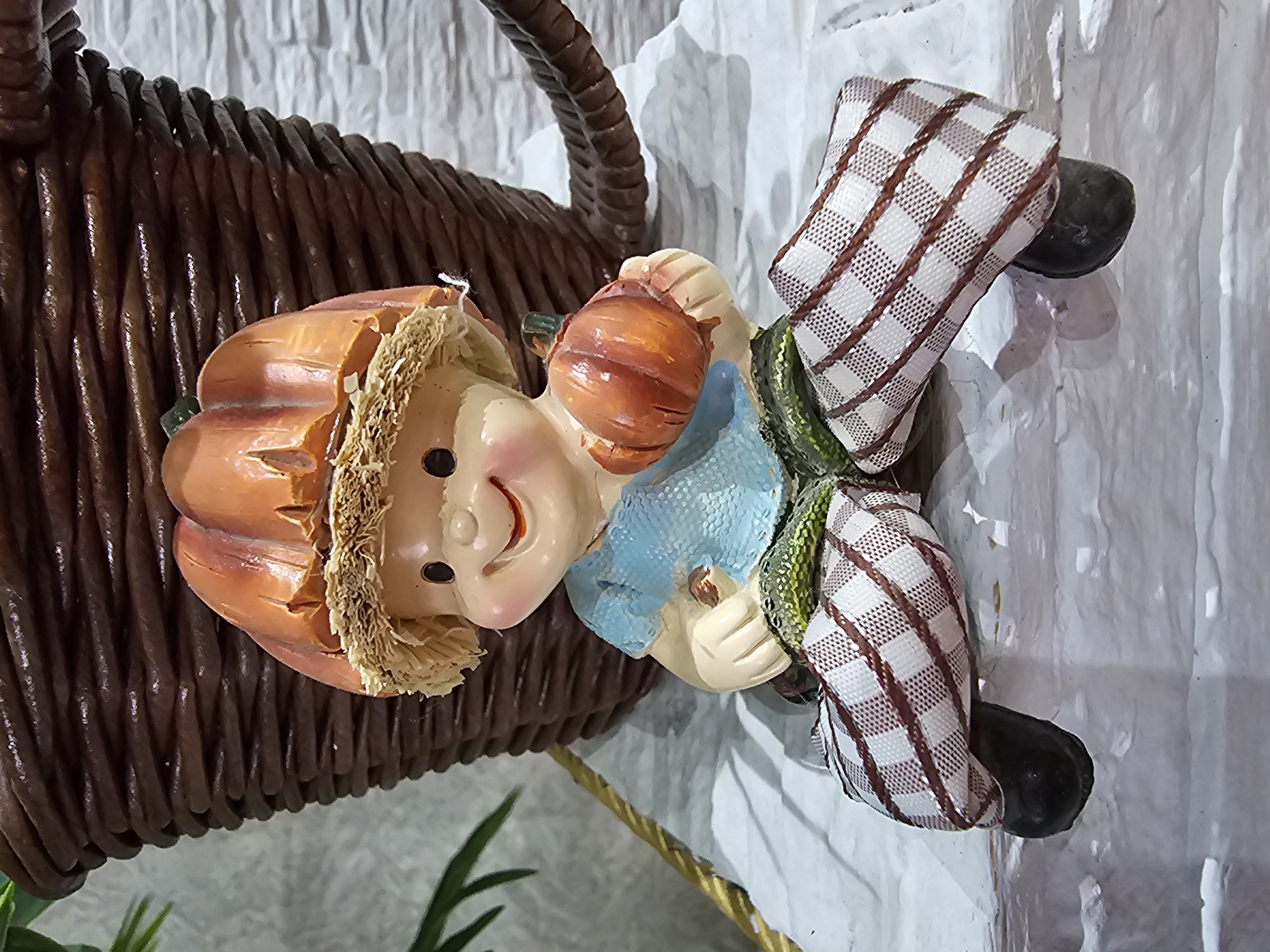 A close-up, eye-level shot features a ceramic figurine of a child wearing a pumpkin-shaped hat, a light blue shirt, and plaid pants. The figurine is seated with its legs crossed, holding a small pumpkin in its right hand. Behind the figurine is a large, dark brown woven basket. The background consists of a white brick-like texture and a blurred green plant on the left. The scene is indoors, on a white surface that appears to be a shelf or ledge. The lighting is soft and diffused. There is no visible indication of the city of Kalinovka, Russia in this image.