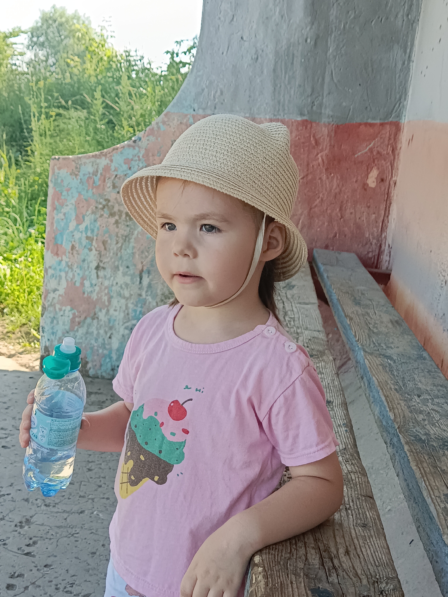 A young girl wearing a straw hat and a pink t-shirt with an ice cream cone print stands outdoors. She is holding a plastic bottle of water with a green cap. The girl is looking to her right with a curious expression.
She appears to be in a rustic setting, possibly near a weathered wooden bench and a wall with peeling paint in shades of pink, blue, and grey. In the background, green foliage suggests an outdoor environment during daylight, likely a warm day given the summer attire. The overall scene evokes a relaxed, possibly casual outing in a rural or semi-rural location within Posad, Russia. There is no visible text of significance.