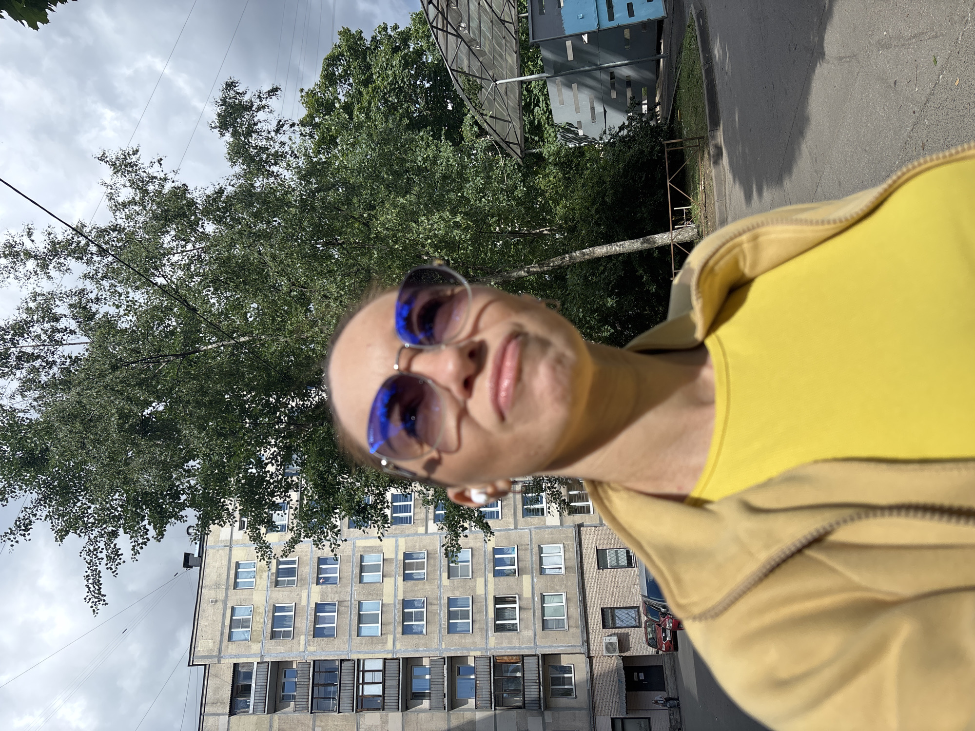 The media file is a selfie taken outdoors, presumably in Saint Petersburg, Russia. The subject is a person wearing sunglasses and a yellow top. The person is looking directly at the camera with a slightly quizzical expression. In the background, a tall, grey apartment building with many windows is visible, along with trees and a cloudy sky. The scene appears to be set during the daytime, with diffused sunlight casting shadows. There are some wires stretching across the frame, likely related to the urban environment. The overall impression is of a casual, everyday moment captured on a cloudy day.