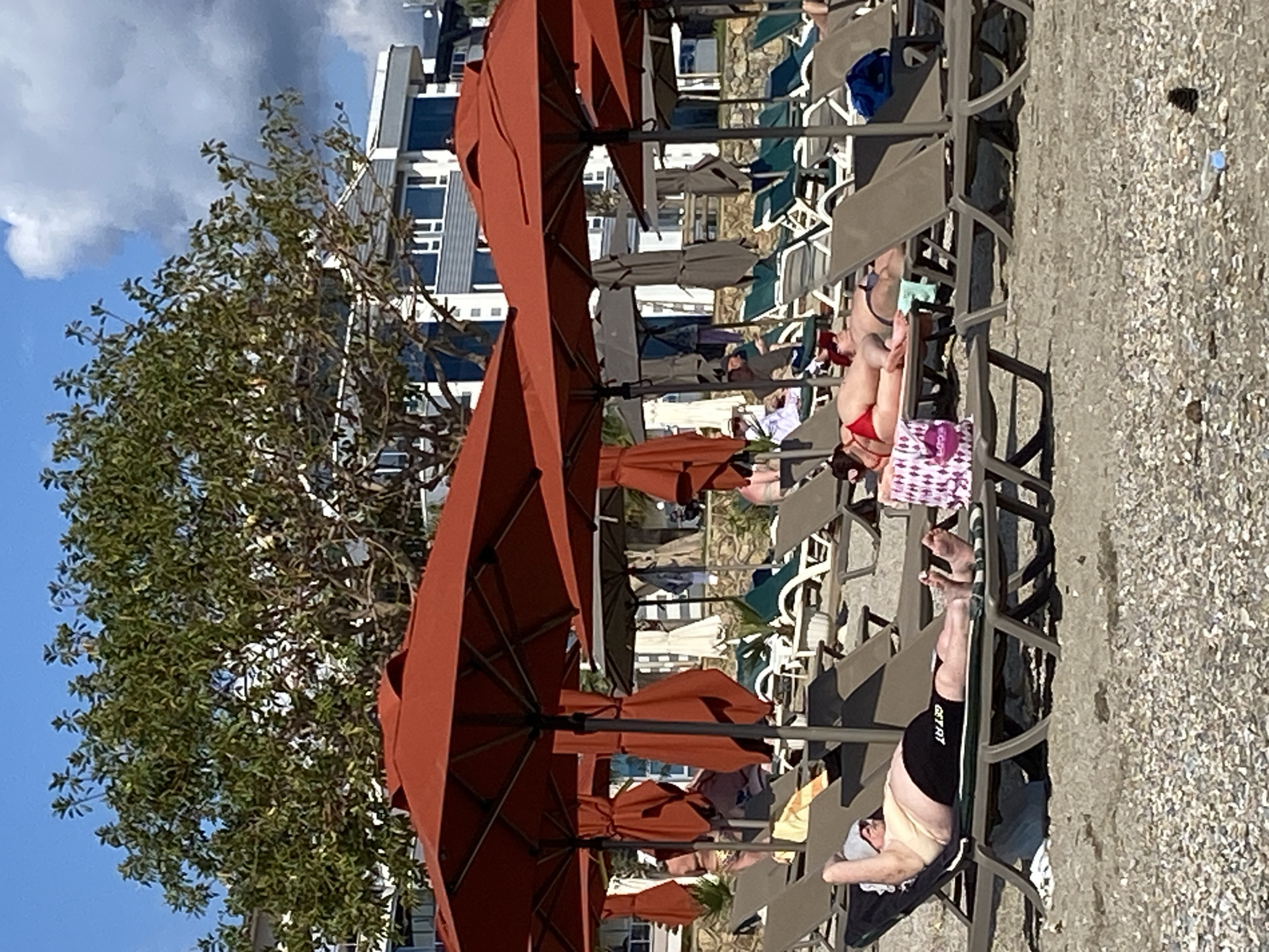 The scene depicts a beach in Alanya, Turkey, filled with sunbathers on lounge chairs under orange parasols. The beach is sandy and textured, interspersed with litter. Buildings can be seen in the background, partially obscured by trees and parasols. People are relaxing and sunbathing on the beach chairs. The overall scene shows a typical sunny day at a resort beach.