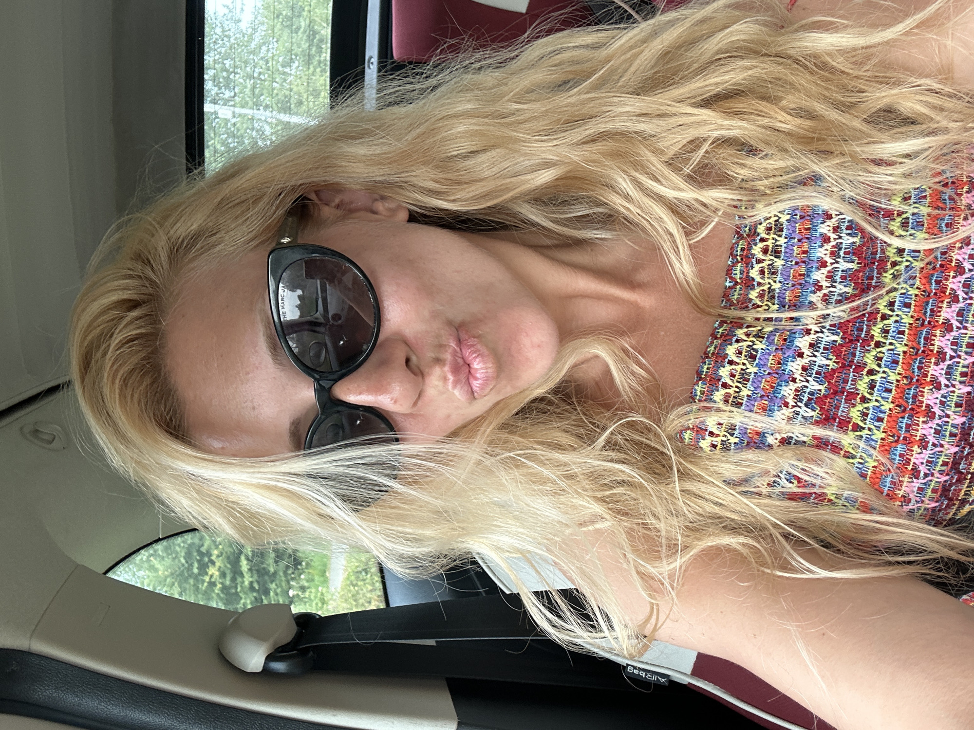 In Cayeux-sur-Mer, France, a blonde woman with long, wavy hair is seen from the chest up, sitting in the passenger seat of a car. She is wearing black, cat-eye sunglasses and a colorful, patterned strapless top. Her lips are pursed as if she is about to kiss, and her hair falls loosely around her face and shoulders. The interior of the car is visible, including a seatbelt and the car's headrest. Through the window, greenery and trees can be seen, suggesting an outdoor setting.