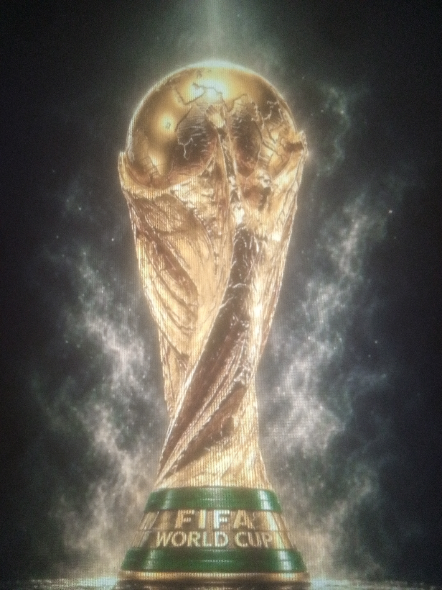 The image displays a close-up, low-angle shot of the FIFA World Cup trophy. The trophy is golden and appears to be made of polished metal, with intricate detailing representing the Earth. It is set against a dark, smoky, or nebulous background with subtle lighting effects that create a dramatic and ethereal atmosphere. The base of the trophy is visible, adorned with the text "FIFA WORLD CUP" in a metallic font. The overall presentation suggests a celebratory or prestigious context, possibly a promotional image or a depiction of victory. The image does not contain any elements that can be used to identify a specific location in Tundun Wada West, Nigeria, or any other geographical area.