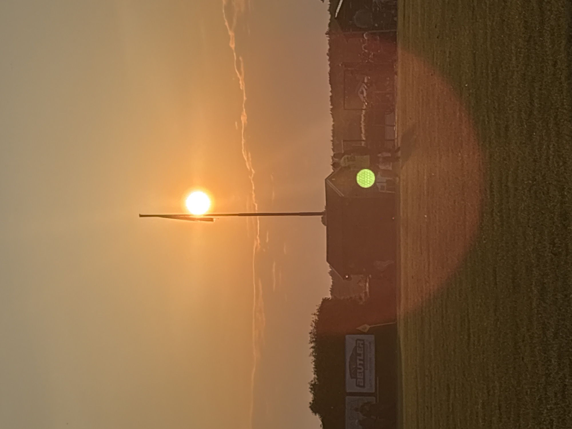 The image depicts a landscape at sunset in Wegberg, Germany. The sun is positioned on the left, slightly obscured by a flag pole. The sky is a gradient of orange and yellow hues. To the right of the flag pole, a building is visible, next to a grassy field. A sign with the word "BEUTLER" is at the edge of the field.