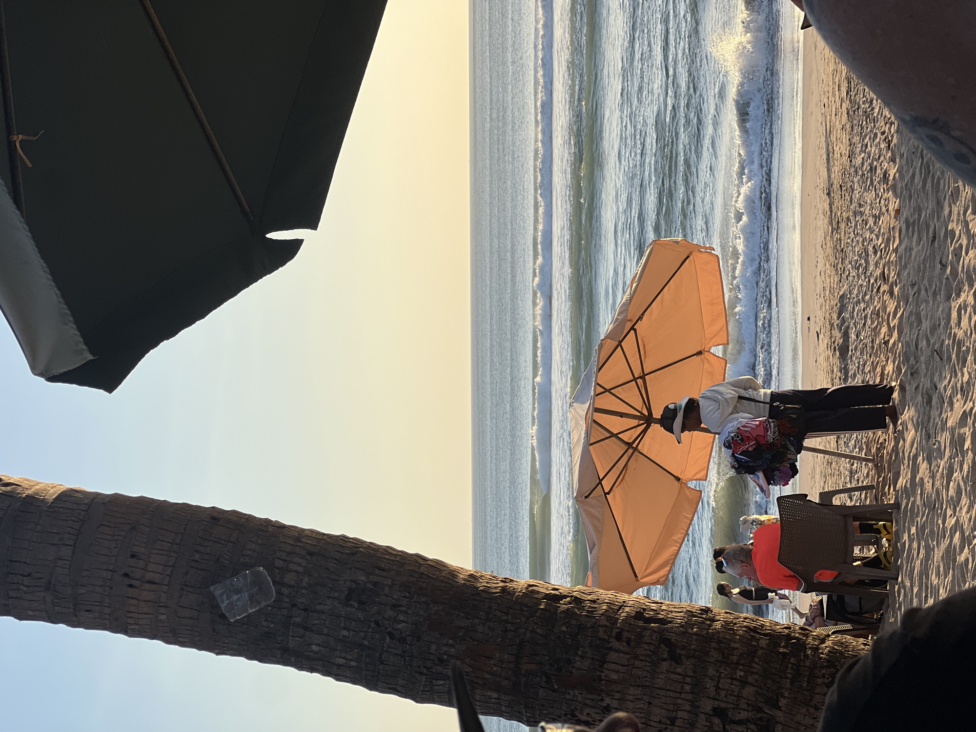 The image depicts a beach scene in Kuta, Indonesia, during what appears to be late afternoon or early evening. In the foreground, a large palm tree trunk dominates the left side of the frame. A dark umbrella partially obscures the upper right portion of the image.
A light orange beach umbrella is set up on the sand, casting a shadow. Beneath this umbrella, a person wearing a white hoodie and a baseball cap stands next to a display of various colorful items, possibly souvenirs or clothing, hanging from a rack. To the left of the umbrella, a person in a red shirt is seated in a brown chair, facing away from the camera. Their legs are visible, wearing yellow shoes.
In the mid-ground, the ocean stretches out to the horizon, with gentle waves breaking on the shore. Several people are visible near the water's edge, some appearing to walk or stand. The beach is covered in sand, with some ripples and footprints visible. The sky is a pale yellow and blue gradient, indicative of a sunset or sunrise. The overall atmosphere is relaxed and tropical.