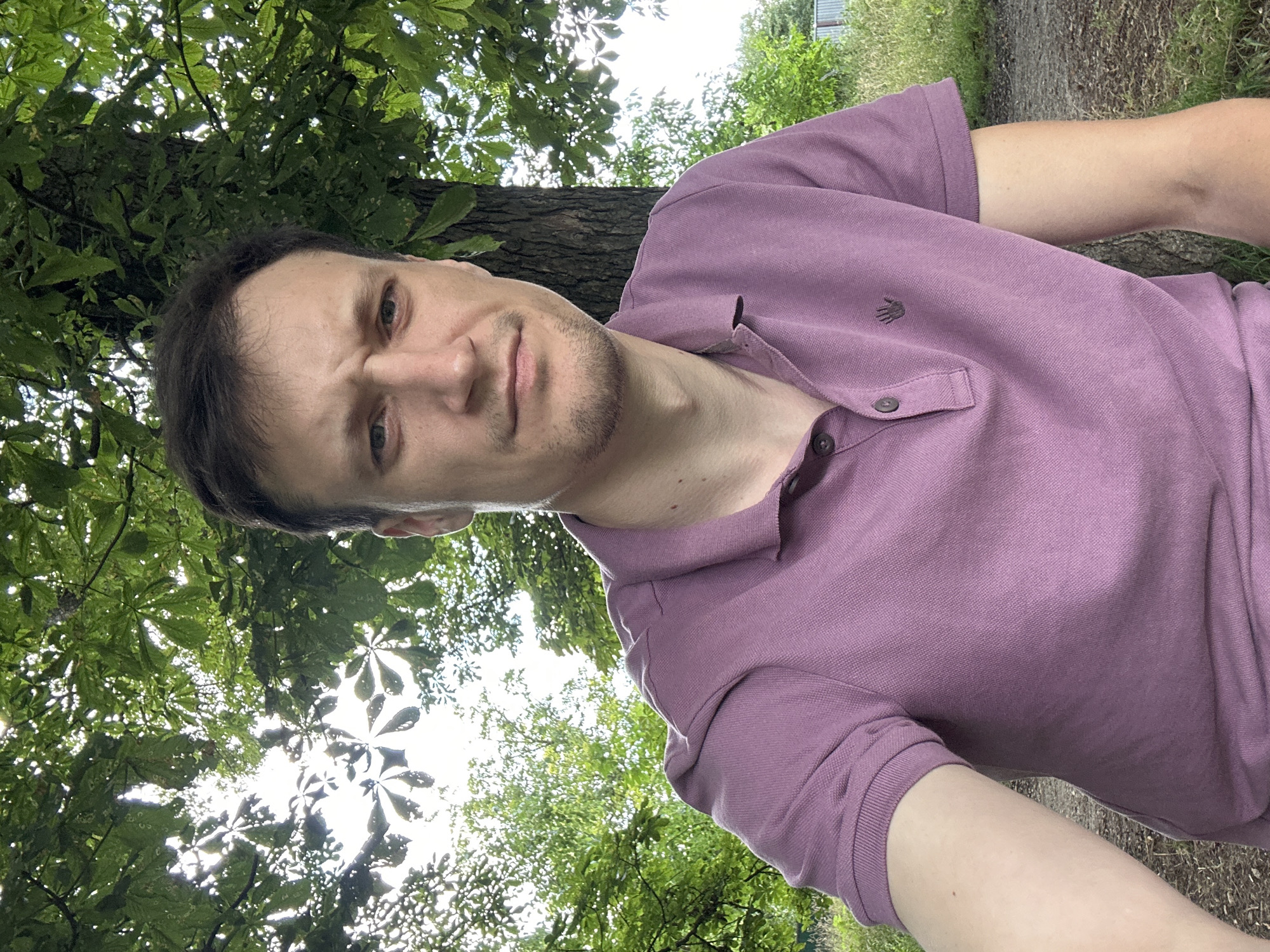 A man with light skin, brown hair, and light blue eyes is pictured outdoors. He is wearing a light purple polo shirt. The man is looking directly at the camera with a serious expression. He has a light beard and stubble. Behind him, a tree trunk is visible on the right side of the image, with green leaves and branches creating a canopy overhead. The background consists of more greenery, including trees and grass, with a hint of a fence on the far right. The lighting suggests it is daytime, possibly overcast or late afternoon due to the diffused sunlight filtering through the leaves. The location is identified as Makiv, Ukraine.