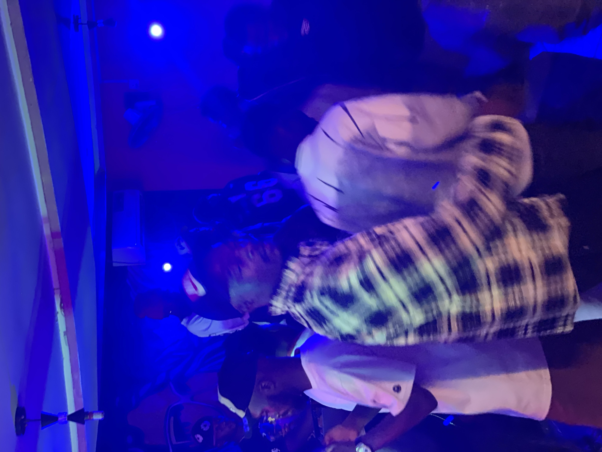 The image depicts a dimly lit, indoor scene that appears to be a nightclub or bar in Durumi, Nigeria. The atmosphere is saturated with vibrant blue lighting, creating a cool and energetic ambiance. Several individuals are visible, their forms slightly blurred due to motion, suggesting a lively environment with dancing or movement.
In the foreground, a person wearing a white t-shirt and a black and white checkered open shirt is the most prominent subject. Their body is angled to the right, and their face is partially obscured. To their left, another individual in a white t-shirt and a black baseball cap is visible from the side. Their head is turned to the left, and they appear to be holding something in their hand.
Further back, other people can be seen, their features less distinct due to the lighting and distance. One person in the background is wearing a black jersey with the number "66" in white. The walls are painted a deep red, contrasting with the dominant blue lighting. A ceiling fan is mounted on the wall to the right, and various light fixtures are suspended from the ceiling. The overall impression is one of a social gathering at night, likely in a popular entertainment venue.