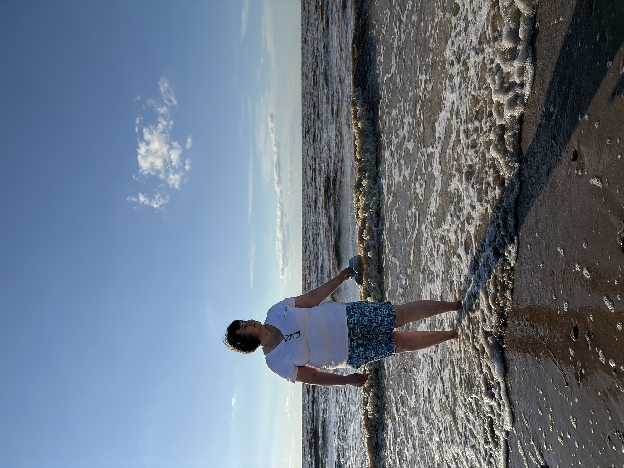 In the city of Port-Katon, Russia, a woman stands in shallow ocean water under a blue sky with a single cloud. She is wearing a white t-shirt and blue patterned shorts, and is holding a pair of light-colored shoes in her right hand. The water is slightly choppy with small waves, and the shoreline is sandy with some foam.
