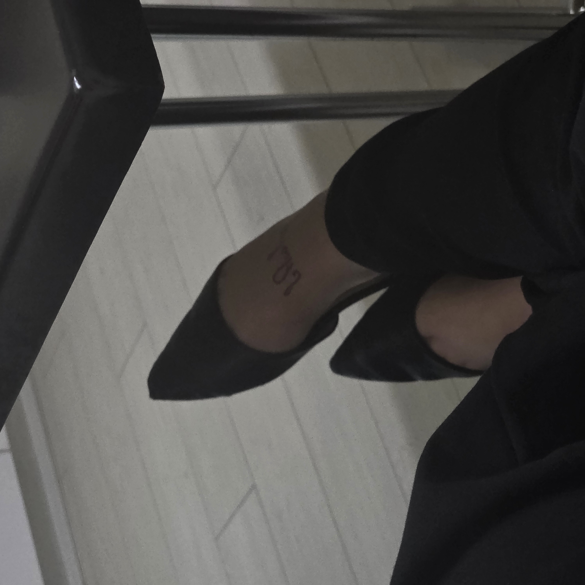 The image is a close-up, top-down view of a person's legs and feet wearing black, pointed-toe shoes. The person has a tattoo on the top of their left foot that appears to read "10.L." The scene is indoors, with a light-colored floor that looks like wood or laminate flooring. A dark, reflective surface, possibly a table or desk, is visible in the upper portion of the image, with metal legs of a chair or table visible on the right. The lighting is dim, creating shadows and a somewhat muted color palette. No specific landmarks or context suggesting a location within Chișinău, Moldova, are present in the image.