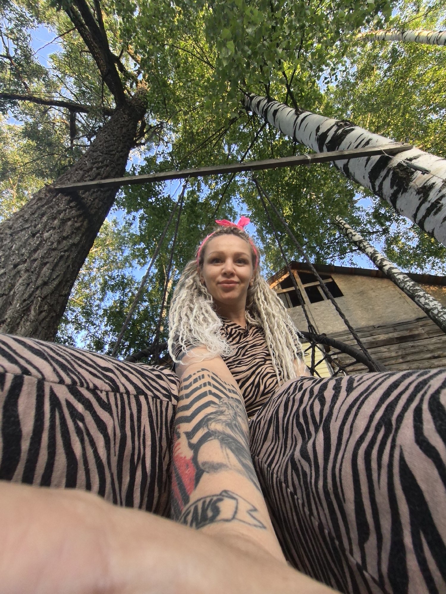 A low-angle view captures a woman sitting on a swing suspended between trees. She has light skin, blonde curly hair, and wears a pink headband. She is dressed in a zebra-striped outfit. A tattoo is visible on her left arm. The swing hangs from a wooden beam attached to two trees; one is a birch tree, and the other has a rough bark. In the background, there is a wooden structure with a visible window, likely a treehouse. The location seems to be outdoors, possibly in the vicinity of Noviy, Russia. The overall scene gives a relaxed, natural feel.