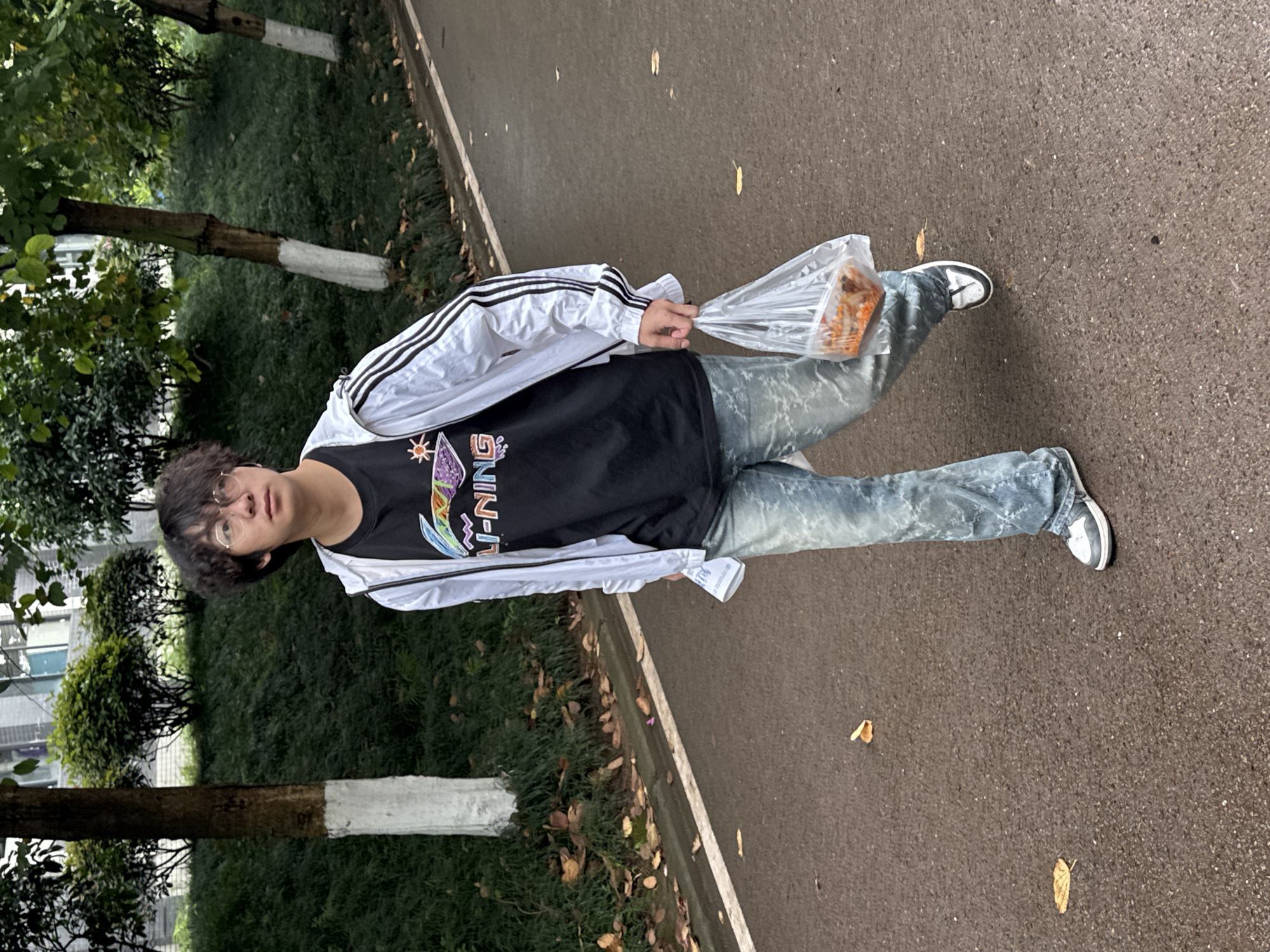 A person is standing on an asphalt pathway with green foliage to the left. The person is wearing glasses, a black t-shirt with colorful graphics, a white jacket with black stripes on the sleeves, and light blue patterned jeans with white and gray shoes. They are holding a clear plastic bag containing something orange. The location is outdoors, likely in Jiangjin, China, based on the user's instruction.