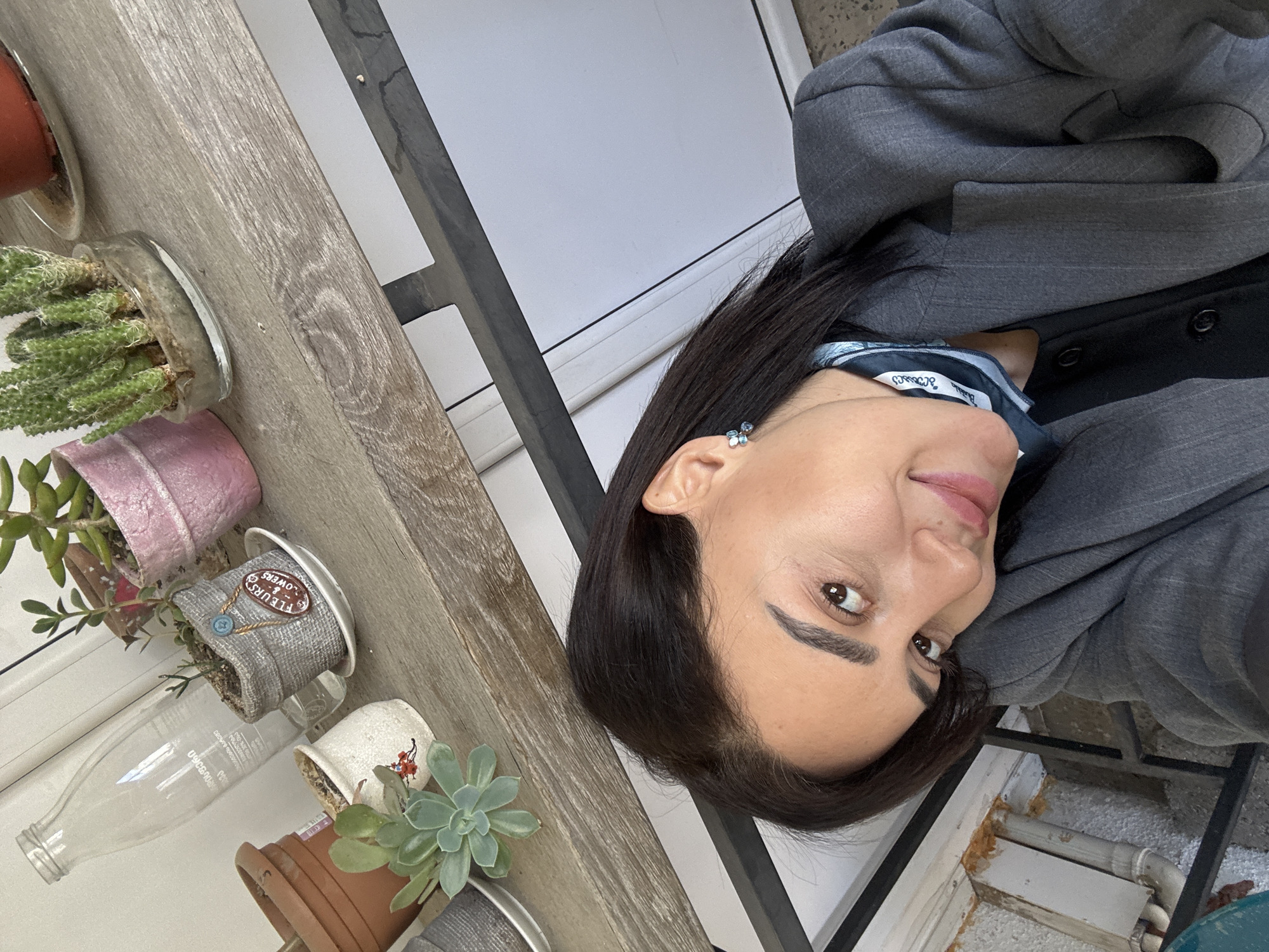 A medium shot captures a woman from the chest up, with her head tilted to the side. She has dark hair, fair skin, and is wearing a gray suit jacket and a patterned blue scarf. Her expression is slightly smiling, and she gazes directly at the camera. An earring is visible on her left ear.
To the left of the woman, a wooden shelf displays a variety of potted plants. The pots range in color and material, including terracotta, painted metal, and fabric-covered containers. Plants in the pots appear to be succulents and cacti.
The background consists of a light gray or white wall, and a black metal frame. Below the woman, a white wall with some pipes.
Based on visual clues and the assumption of location in Tbilisi, Georgia, it is possible that the scene is on a balcony or in a garden area attached to an apartment building in the city, but that cannot be said with certainty.