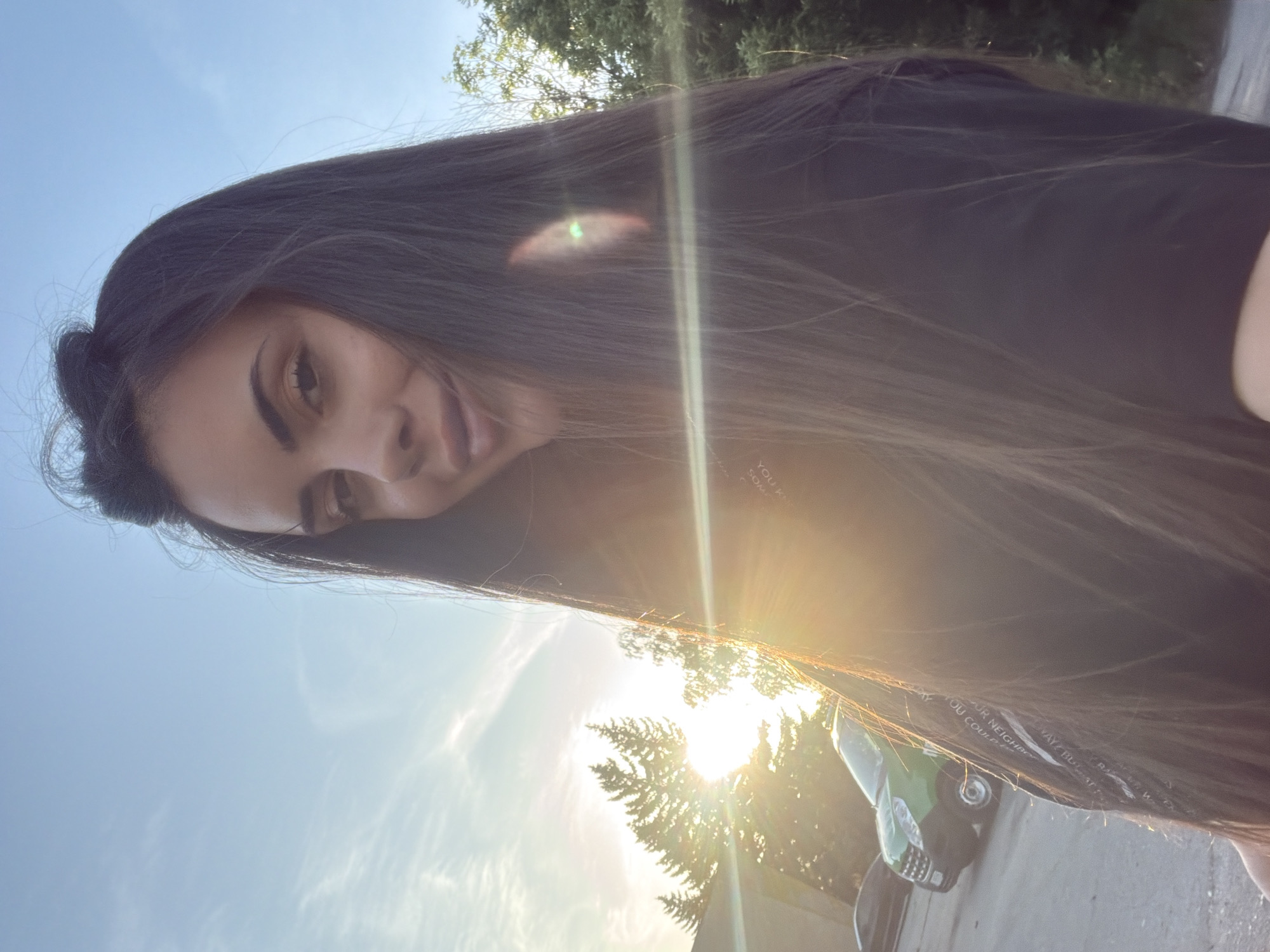 A young woman with long, dark hair poses for a selfie outdoors. She is wearing a black t-shirt with white text. The photo is taken looking upwards, with the sky and treetops visible in the background. A green vehicle is parked nearby. The location appears to be an urban environment, though specific details to pinpoint it within Sofia, Bulgaria, are not discernible from the image.