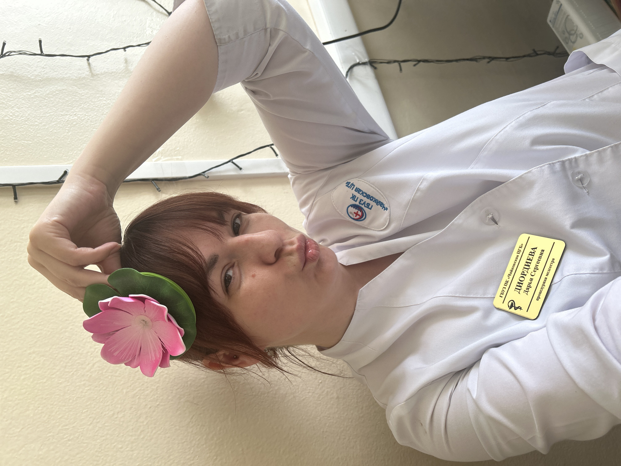 The photograph features a woman with reddish-brown hair, wearing a white medical coat. She has a pink flower and a green leaf accessory in her hair, near her ear. Her lips are pursed. A name badge is visible on her coat, which reads "ДИОРДИЕВА Дарья Сергеевна" in Russian, and indicates her role as a "процедурная медсестра" (procedural nurse) at "ГБУЗ ПК «Чайковская ЦГБ»". This refers to a medical institution in Chaikovsky, Russia. A logo of the institution is visible near the collar. The background appears to be a light-colored wall with a string of fairy lights. The image is taken from a low angle.