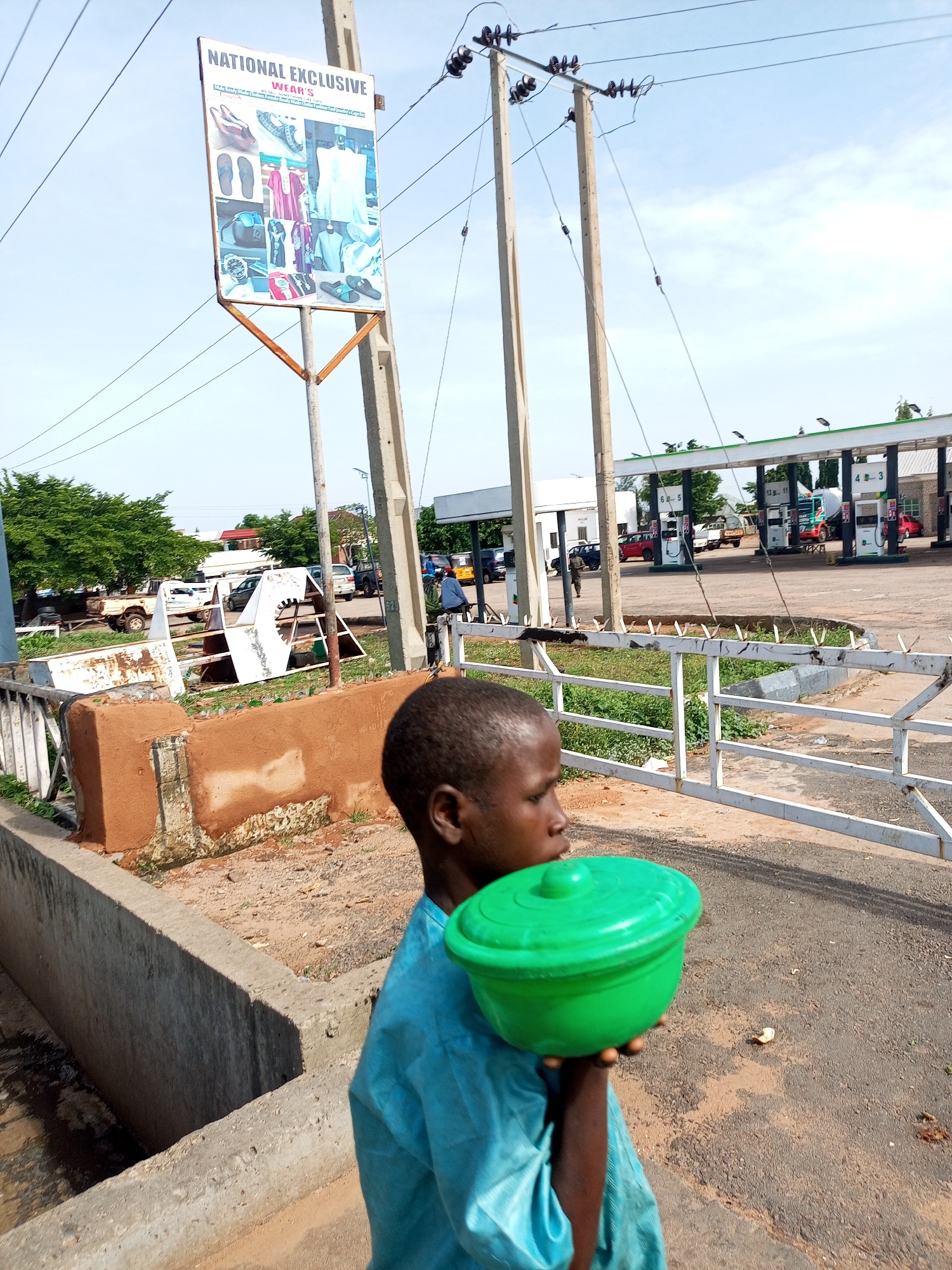 A young Black boy, wearing a light blue tunic, holds a green lidded bowl in his right hand in the foreground. He is facing to the right and looking down. Behind him, a concrete structure with a rough, reddish-brown plaster finish is visible. A white metal fence runs along the top of this structure. In the background, a gas station with multiple pumps and canopies is visible under a partly cloudy sky. Several cars and trucks are parked at the gas station. Power lines crisscross the sky. A sign for "NATIONAL EXCLUSIVE WEAR'S" is mounted on a pole to the left of the frame, displaying images of clothing and footwear. The scene appears to be an outdoor urban environment in Tempure, Nigeria.
