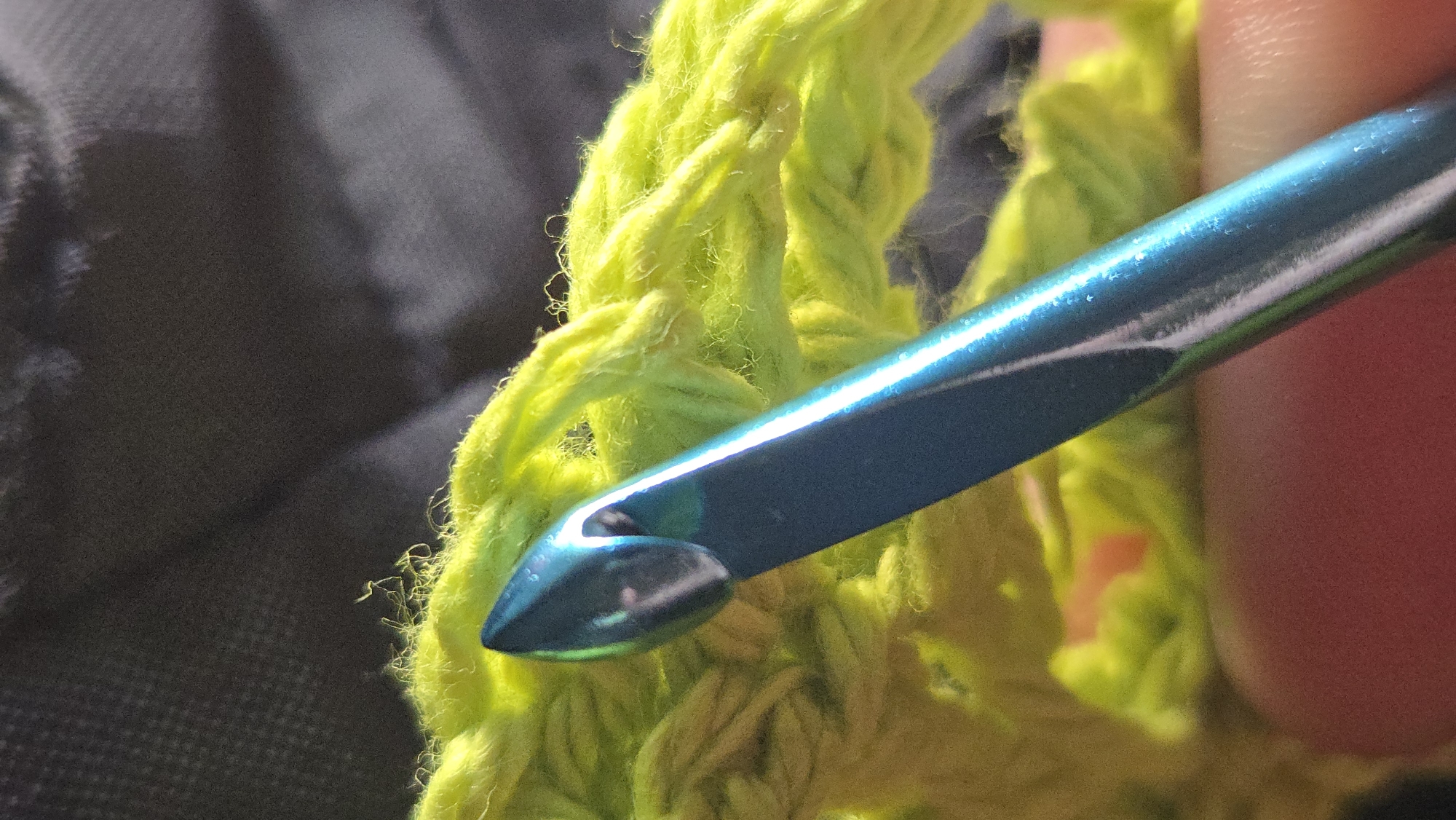 A close-up photograph shows a teal-colored crochet hook in the process of working a stitch with bright green yarn. The yarn appears to be fluffy and somewhat loosely spun. A portion of a hand can be seen holding the hook. The background is out of focus and consists of a dark gray material, possibly clothing or a carrying bag. The image does not offer information about the location. Thus, it is not possible to determine if it was taken in Lompoc, United States of America.