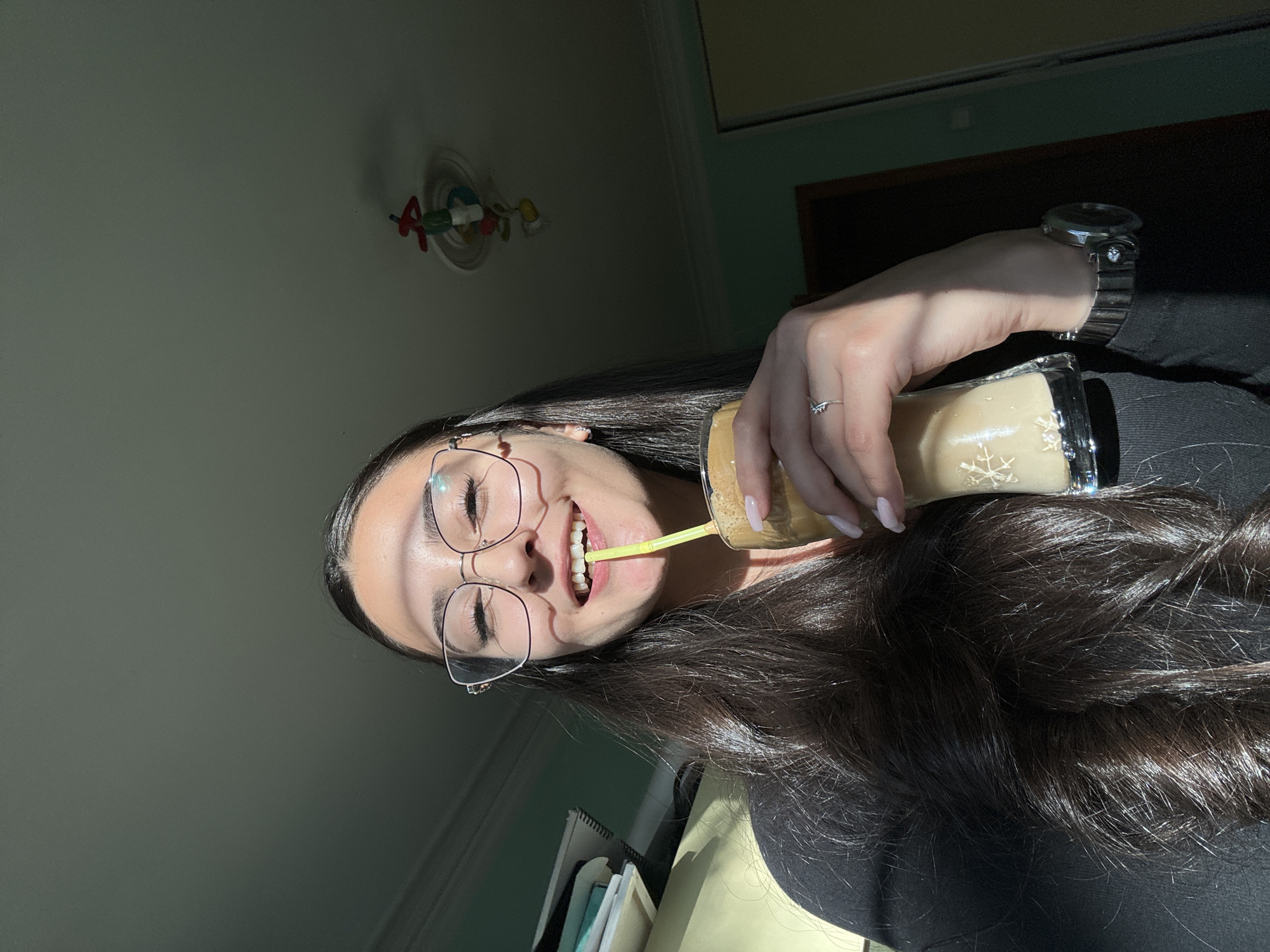 A young woman wearing glasses is drinking from a glass through a yellow straw. She is wearing a dark top and a watch on her left wrist. Her hair is long and dark. The glass has a snowflake design. The location appears to be indoors with a ceiling fixture visible in the background. There is no indication of Dupnitsa, Bulgaria.